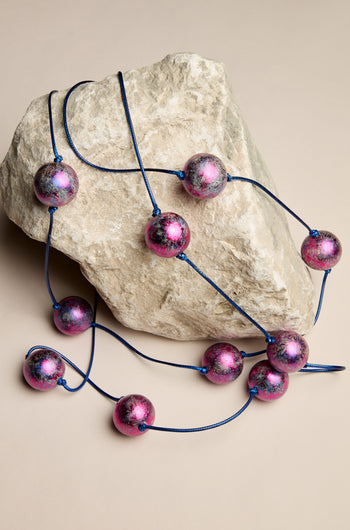 The Metallic Planets Necklace, adorned with large, shiny pink and purple beads on a blue cord, gracefully drapes over a light-colored rock, exuding cosmic elegance.