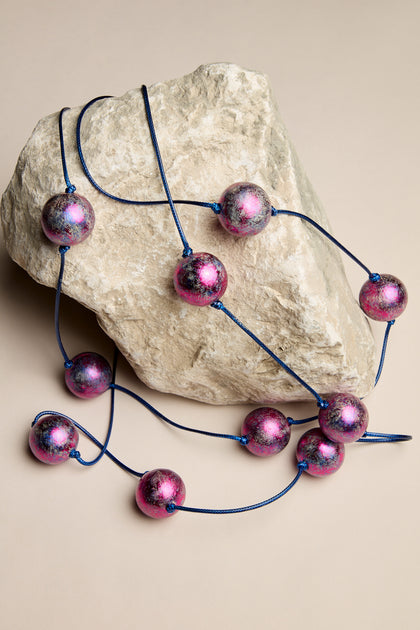 The Metallic Planets Necklace, adorned with large, shiny pink and purple beads on a blue cord, gracefully drapes over a light-colored rock, exuding cosmic elegance.