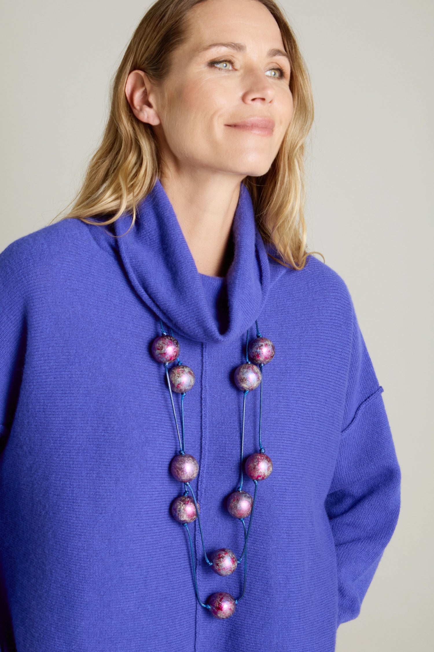 A woman in a blue sweater with a cowl neck smiles, showcasing the Metallic Planets Necklace adorned with large purple and pink beads.