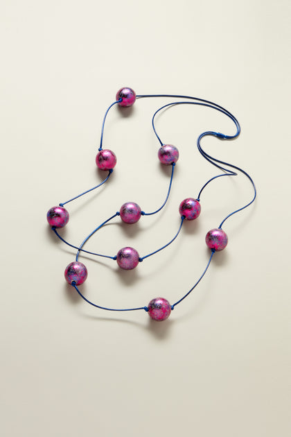 The Metallic Planets Necklace, with its large, round, shiny pink beads strung on a dark blue cord and arranged in a loose, coiled manner, exudes cosmic elegance.