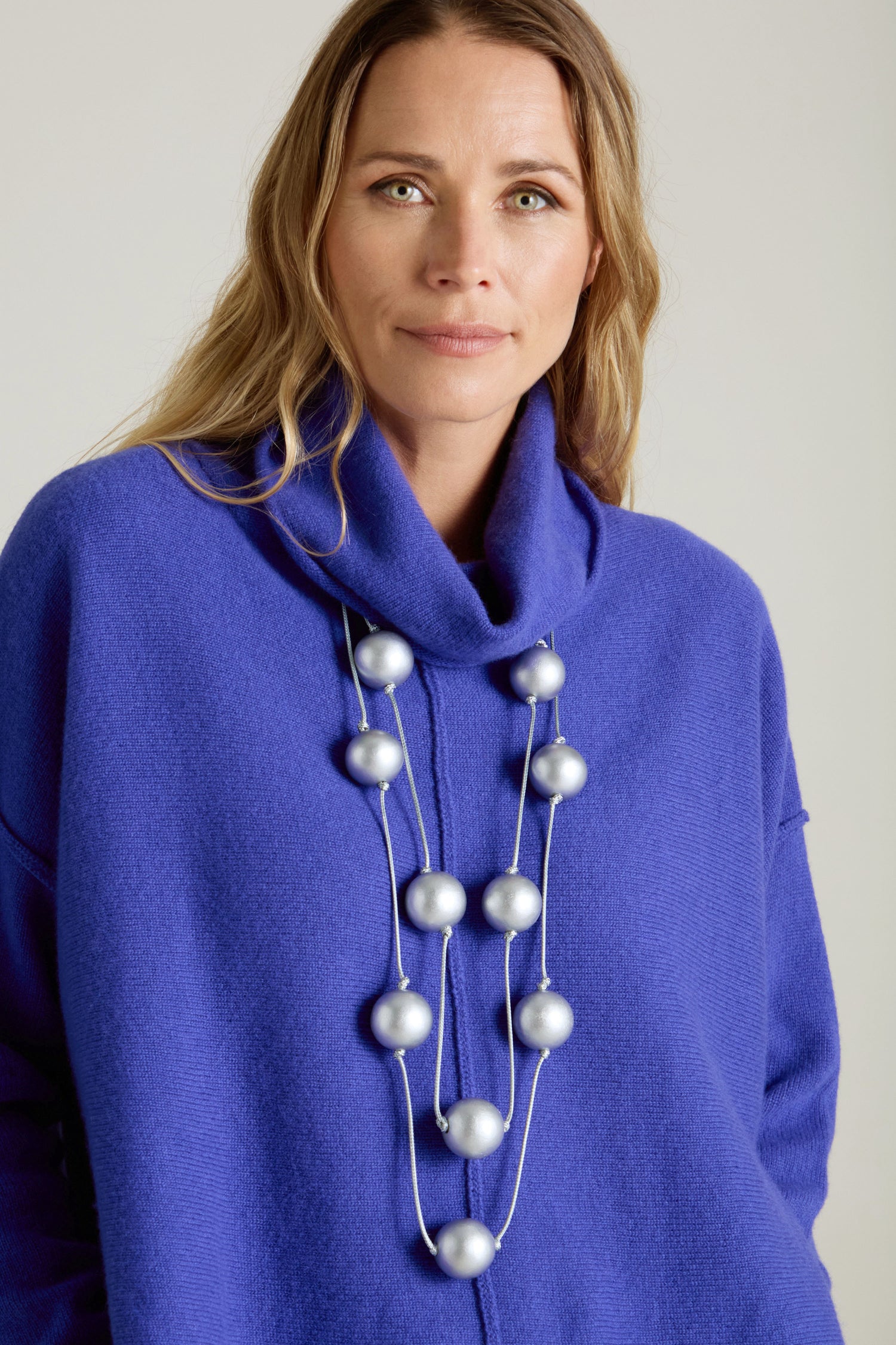 A person wearing a blue sweater and the Metallic Planets Necklace, adorned with large silver beads, gazes at the camera against a neutral background, exuding cosmic elegance.