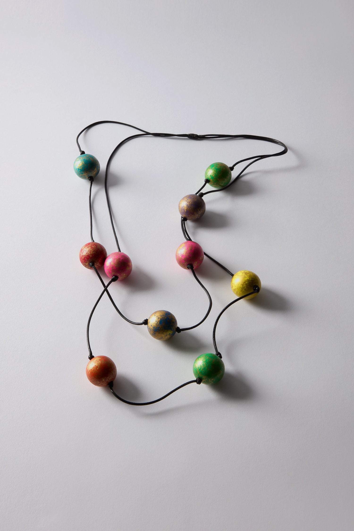 On a light gray surface, the Metallic Planets Necklace stands out as a vibrant statement piece with multicolored spherical beads on a black string, resembling metallic planets.