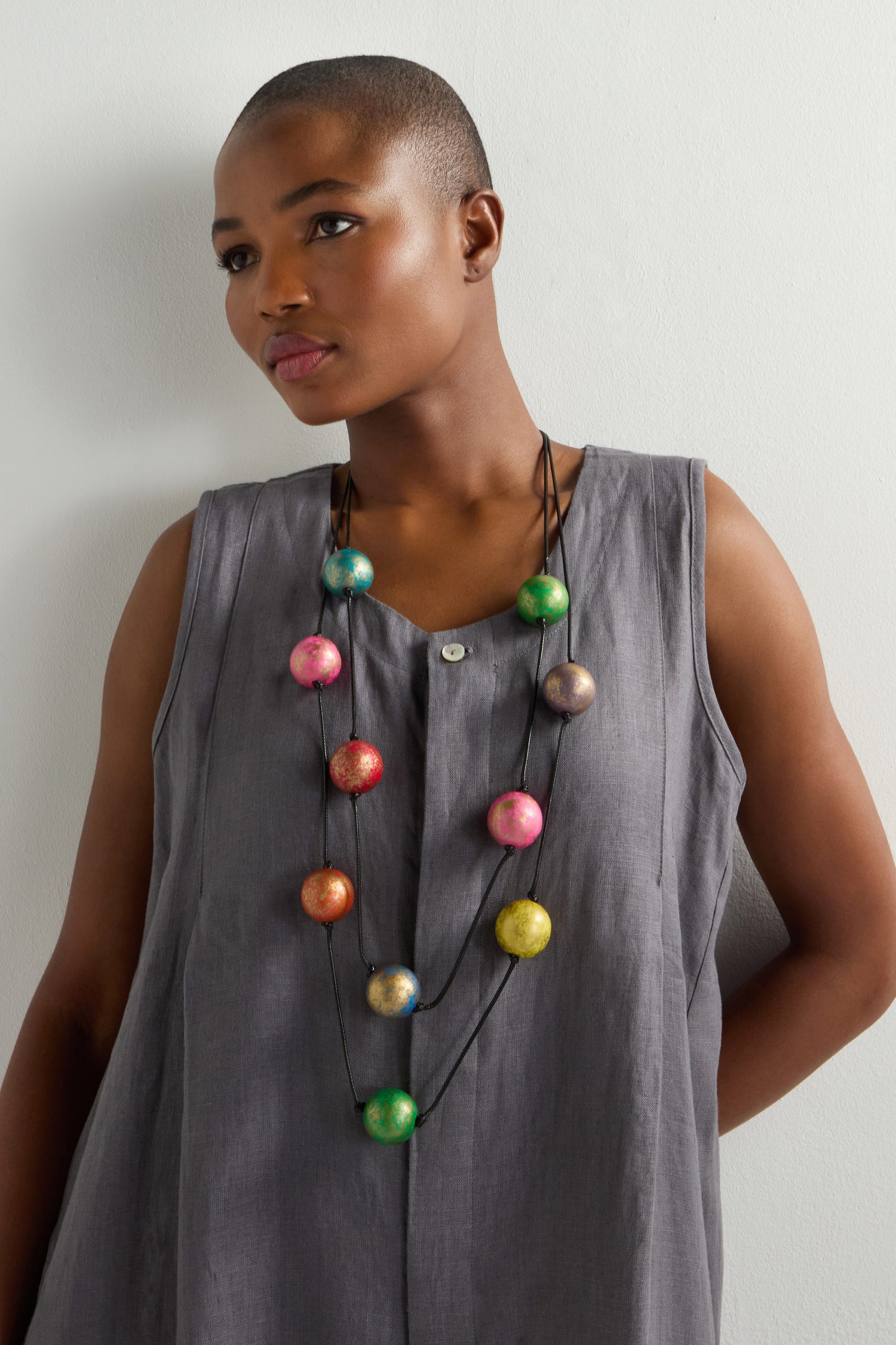 A person in a sleeveless gray dress and vibrant statement piece stands against a plain white wall, wearing the Metallic Planets Necklace adorned with large, colorful celestial-like beads.