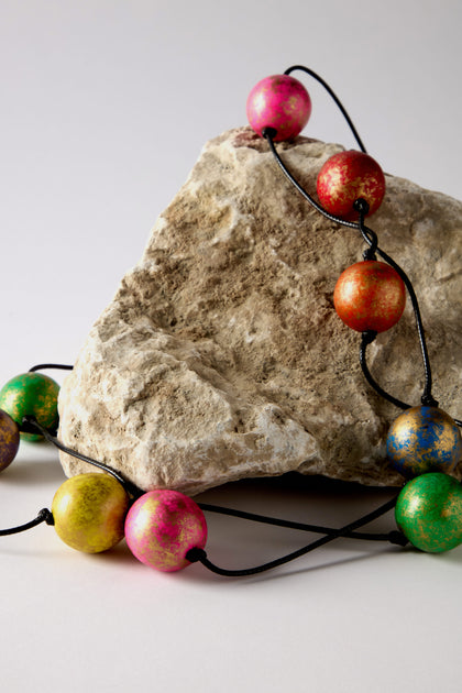 The Metallic Planets Necklace is a vibrant statement piece featuring multicolored round beads draped elegantly over a large, rough rock set against a plain background.