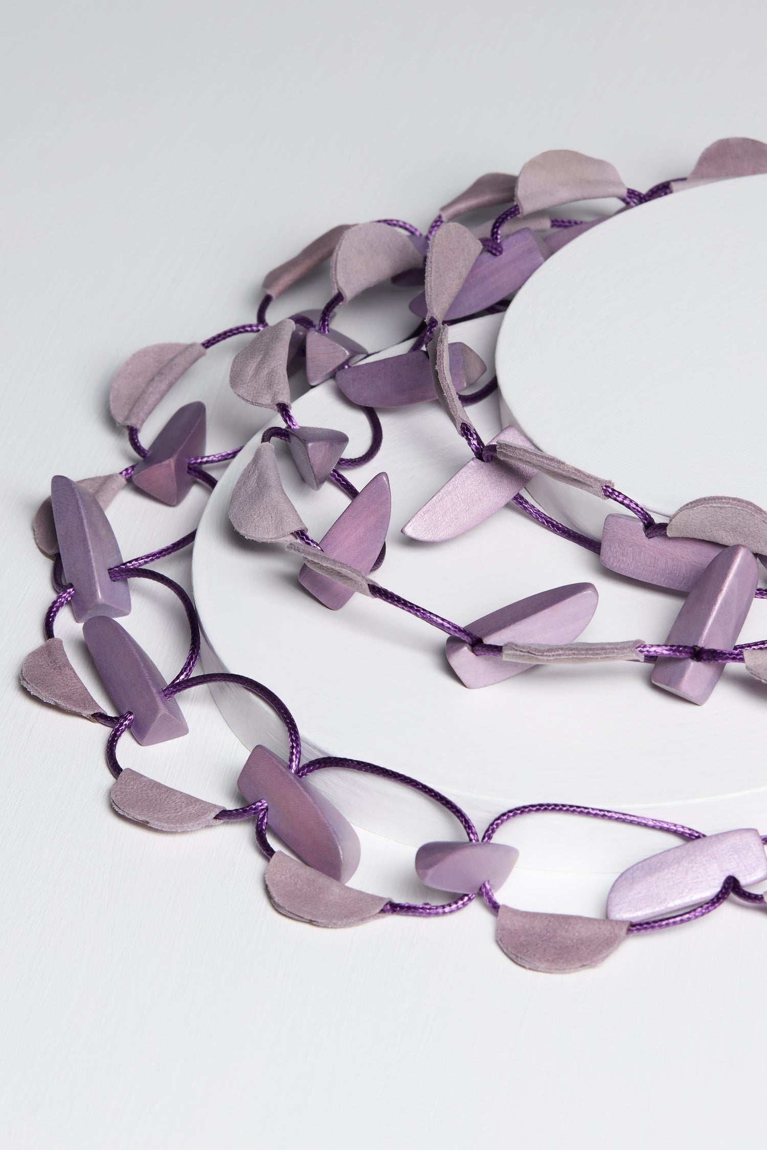 A close-up of the Metallic Shapes Necklace on a white surface, showcasing its purple and lavender irregularly shaped beads. The beads are connected by a soft cord, enhancing the necklace's delicate appeal.