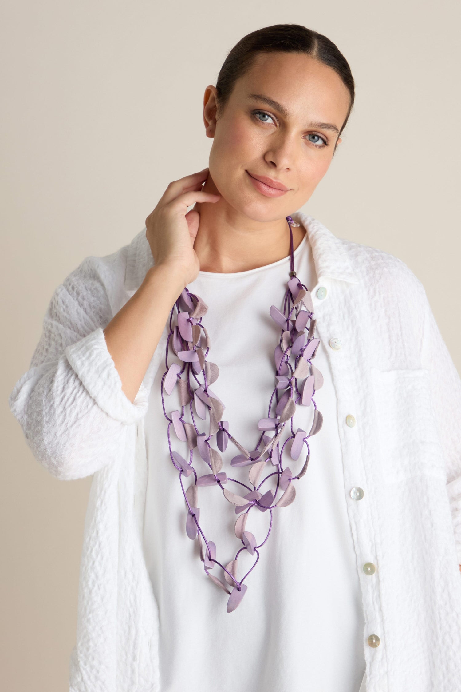 A person wearing a white jacket and white top showcases the Metallic Shapes Necklace, featuring long, purple wooden shapes on a soft cord, while holding their neck with one hand against a plain background.