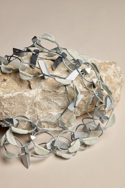 The Metallic Shapes Necklace, featuring interlocking gray and silver metal pieces complemented by lilac wooden shapes, is showcased on a large rock against a beige background.