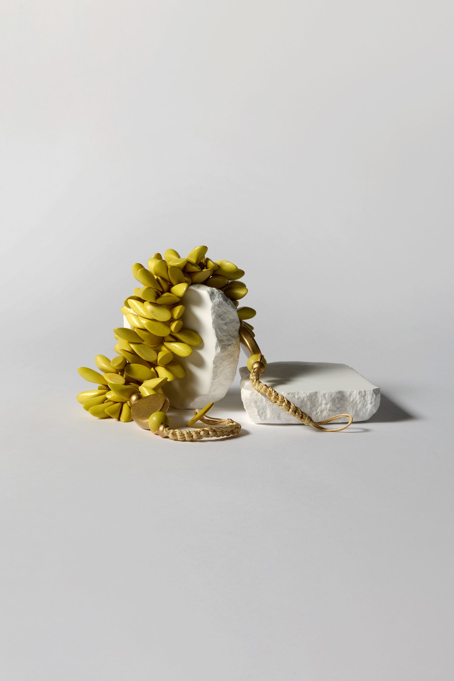 The Citrus Wooden Shapes Necklace features sculptural yellow elongated elements resembling leaves on a white irregular base, evoking a handcrafted aesthetic. Situated on a flat surface with a minimalistic background, it's truly a statement piece.