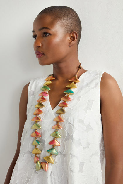 Against a plain white backdrop, a person with a shaved head wears the Metallic Triangles Necklace. Their textured white sleeveless top complements this colorful, cone-shaped bead necklace, exuding sculptural elegance with its iridescent details.