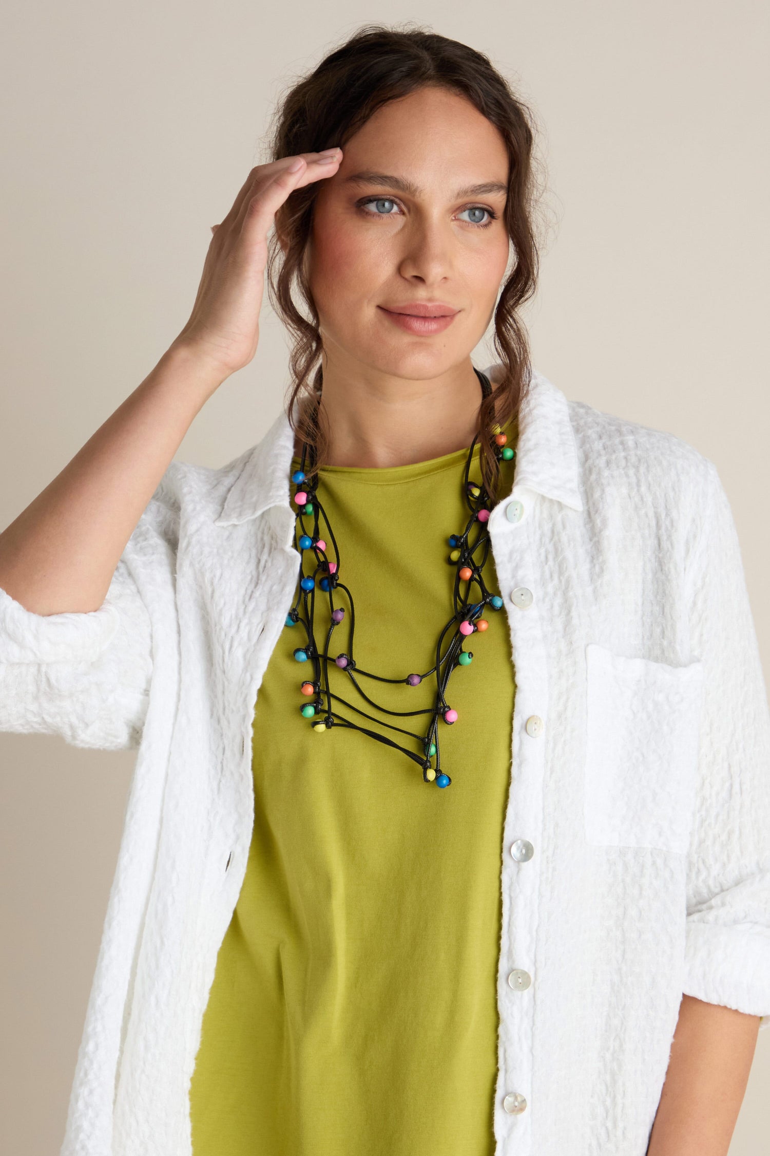 A woman with long brown hair wearing a white button-up shirt over a green top, accessorized with the unique Solar System Necklace featuring multicoloured wooden beads, stands against a plain background. She’s touching her head with one hand.