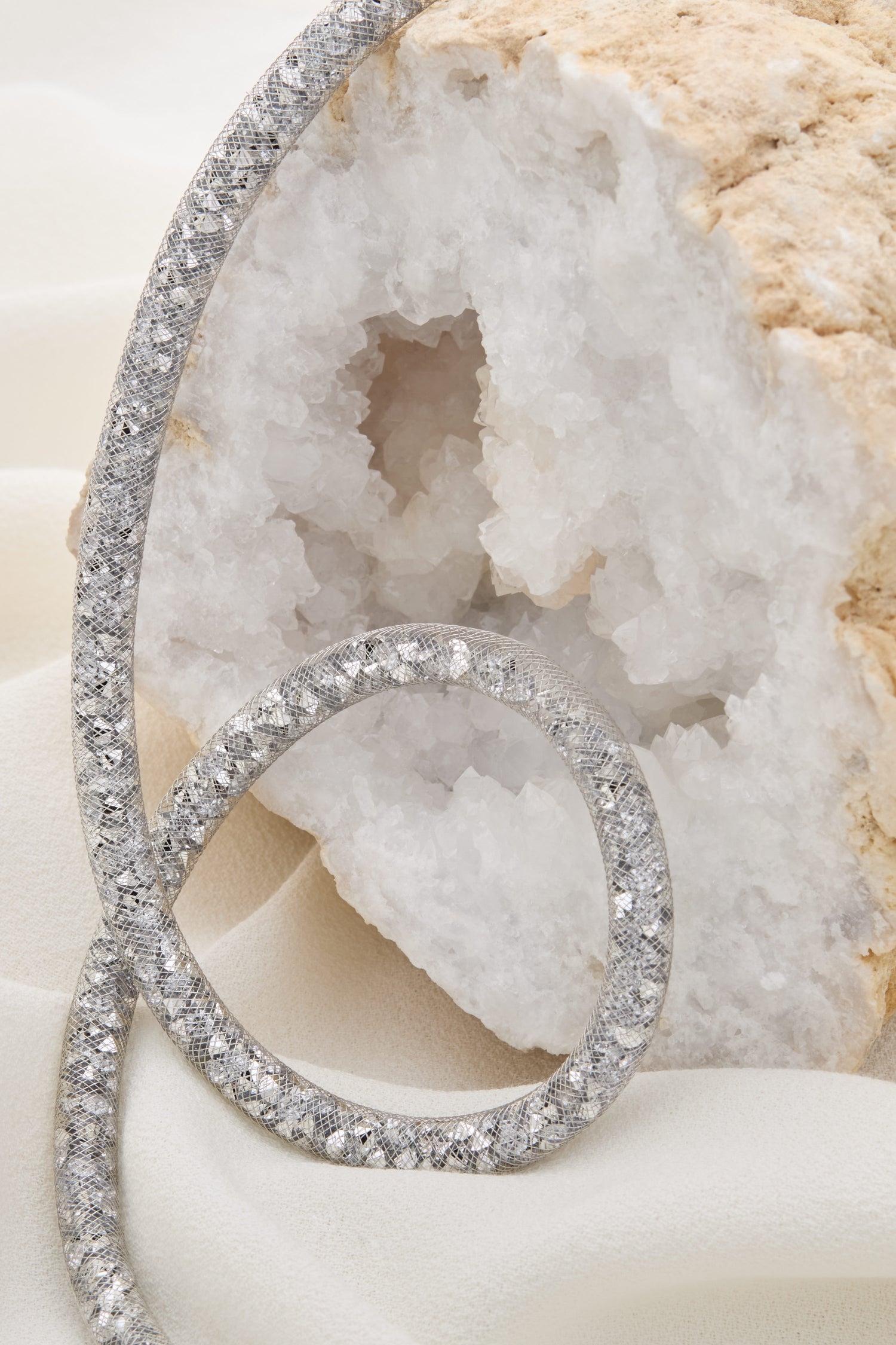 A sparkly, silver textured bracelet is coiled beside a white geode on a light fabric background, complemented by the Long Diamante Necklace featuring an elongated silhouette adorned with shimmering stones.