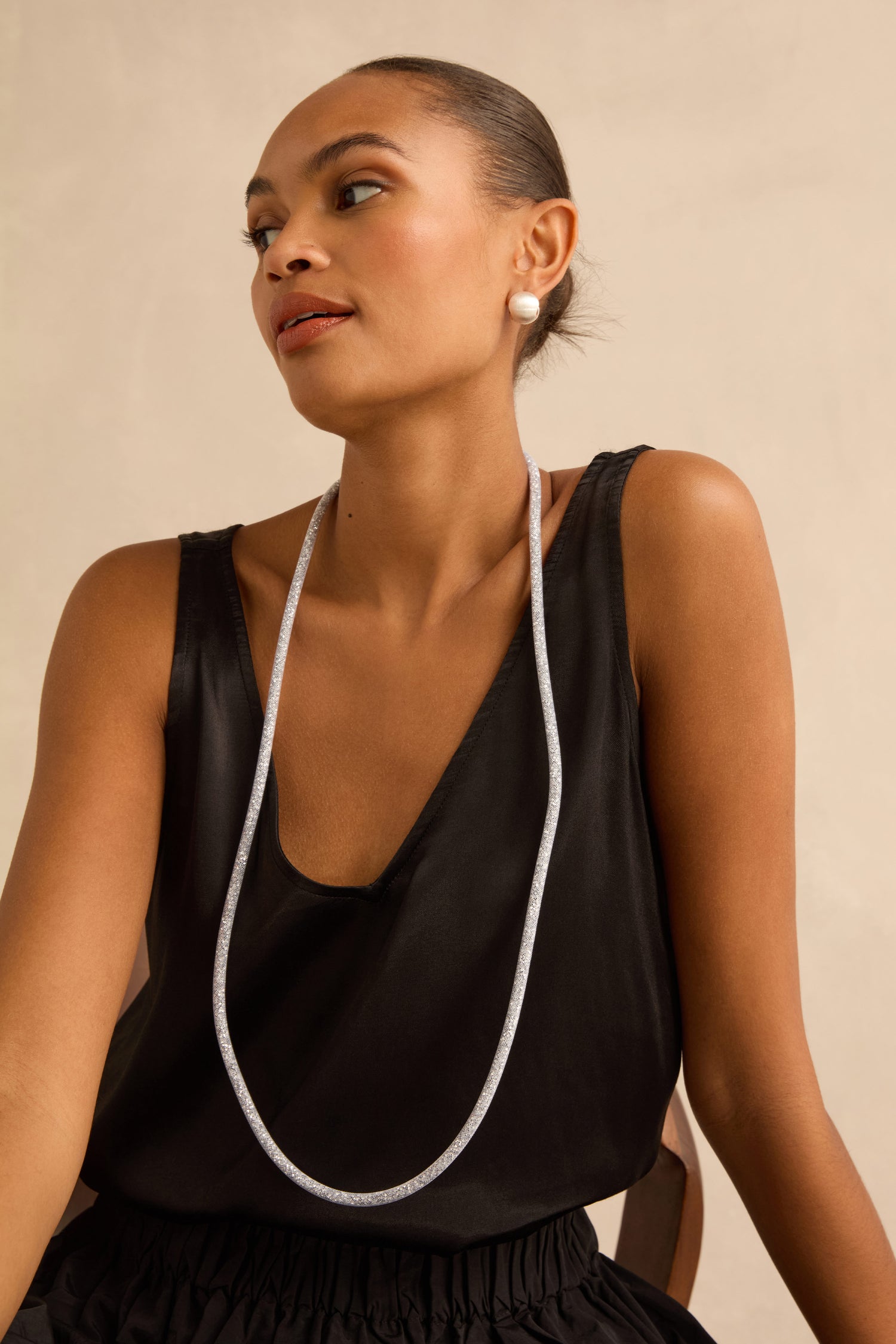 A person wearing a Long Silver Diamante Necklace, styled as a long, slender silver rope, and a pearl stud earring.