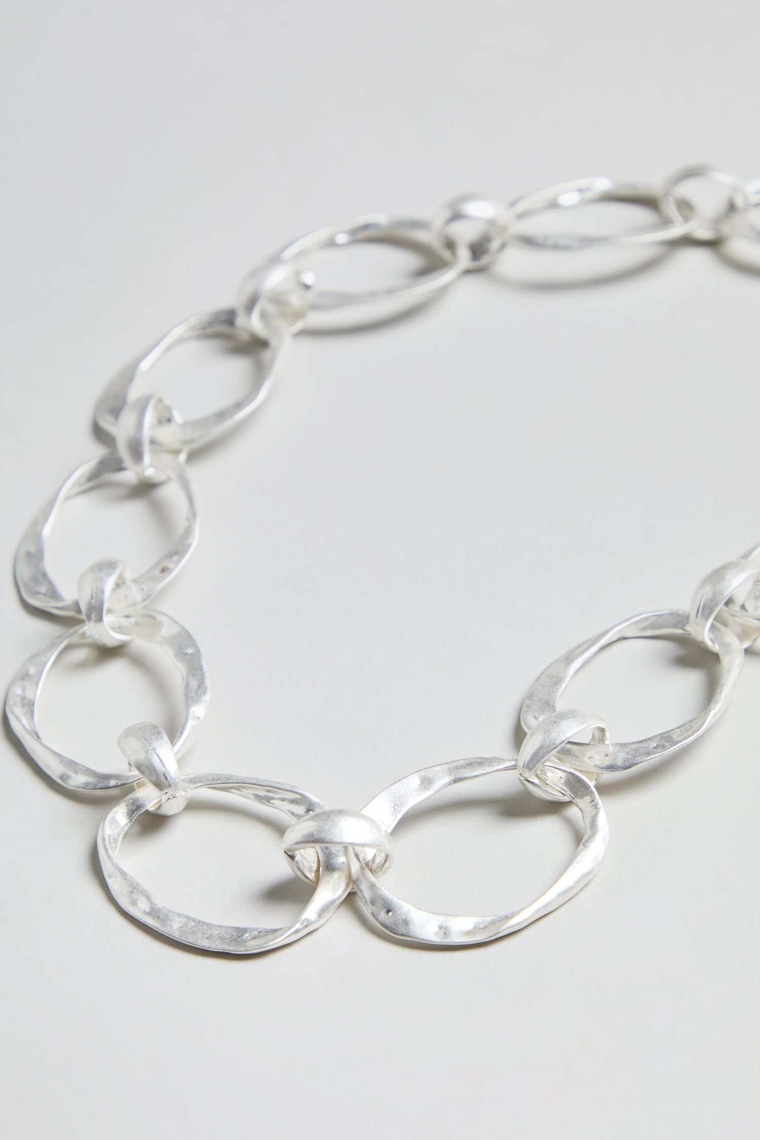 The Hammered Silver Hoop Necklace, with its large, hammered hoop links and silver plating, is displayed flat on a light gray surface.