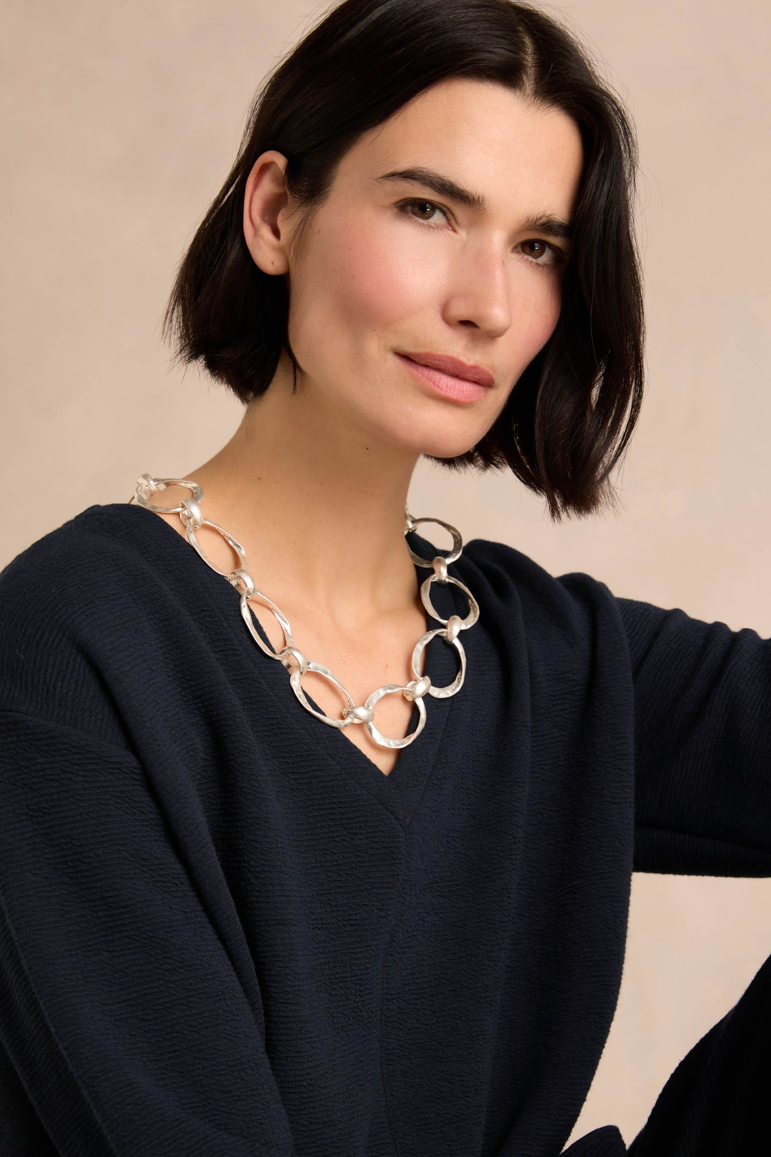 A woman with short dark hair, dressed in a navy sweater, showcases the Hammered Silver Hoop Necklace—a chunky, silver-plated accessory with hammered hoop links—as she poses against a neutral background.