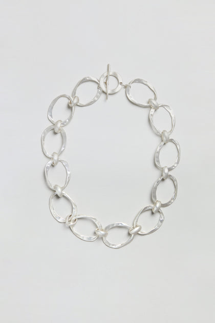 The Hammered Silver Hoop Necklace showcases large, irregular oval links with a hammered finish, joined by smaller round connectors, and is elegantly presented against a plain white background.