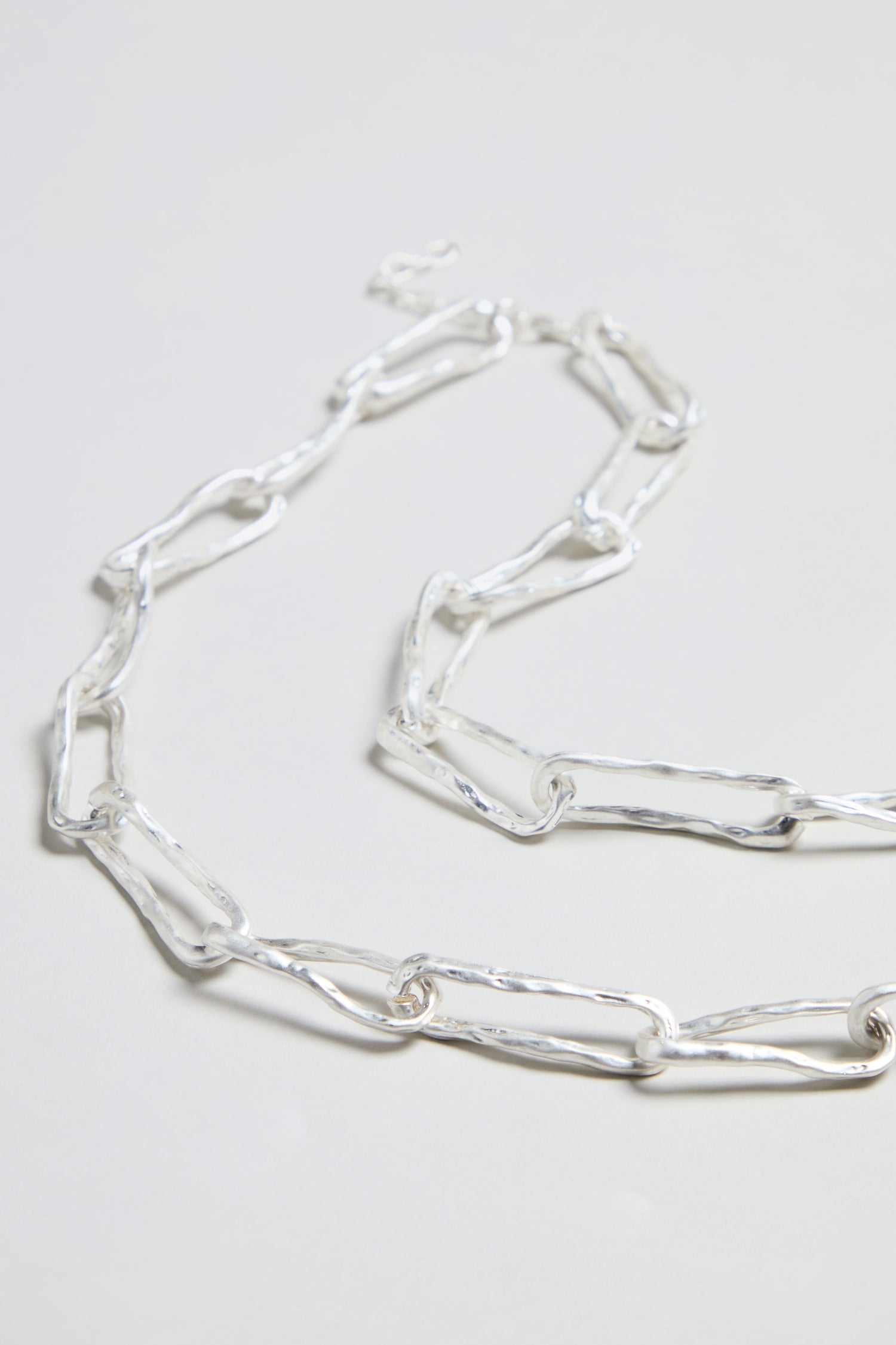 A close-up of the Long Silver Links Necklace arranged on a light grey surface, featuring elongated, irregular silver links and highlighting its unique textured chain.