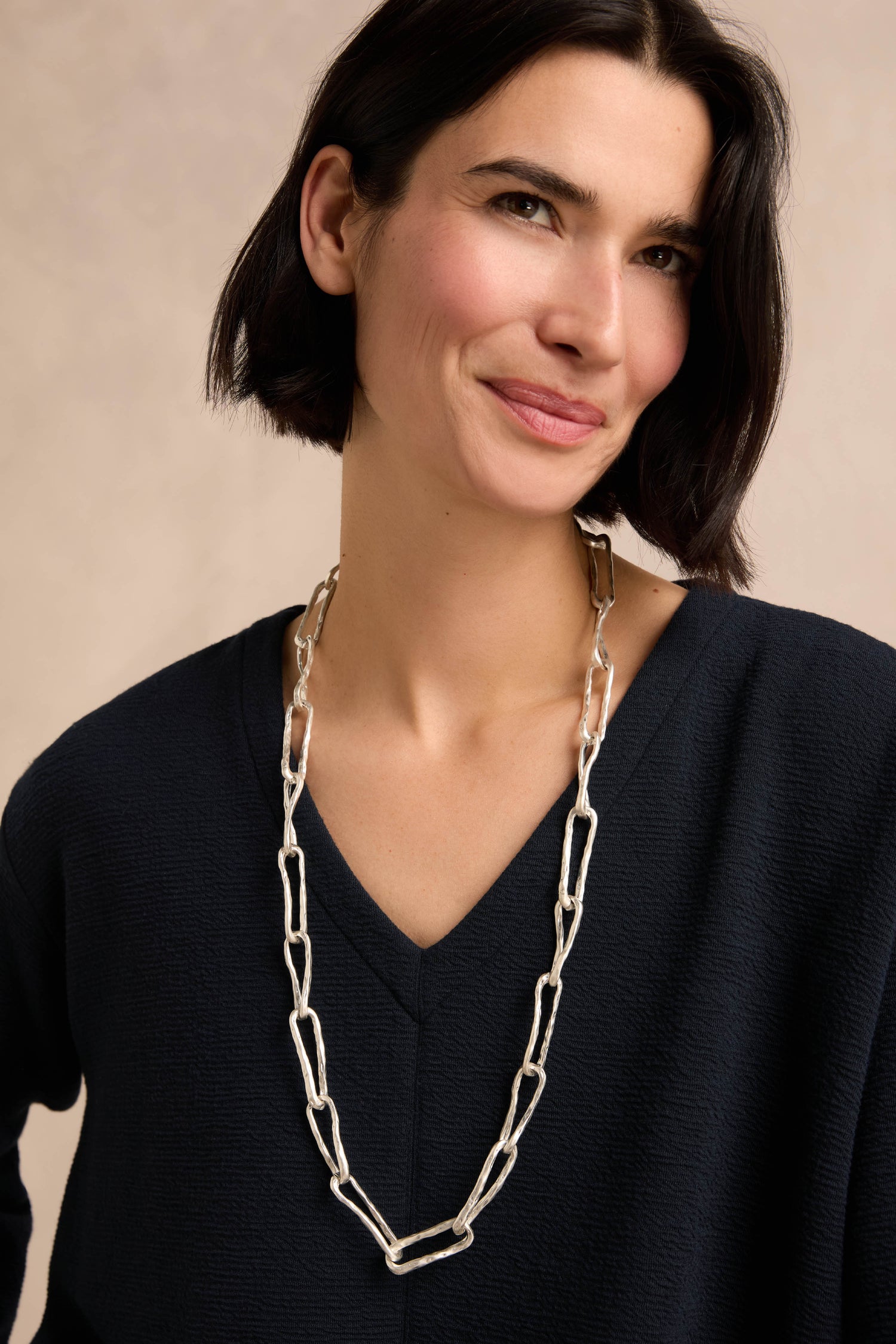 A woman with short dark hair wears a navy V-neck top and the Long Silver Links Necklace—a long, textured chain—while standing against a neutral background.