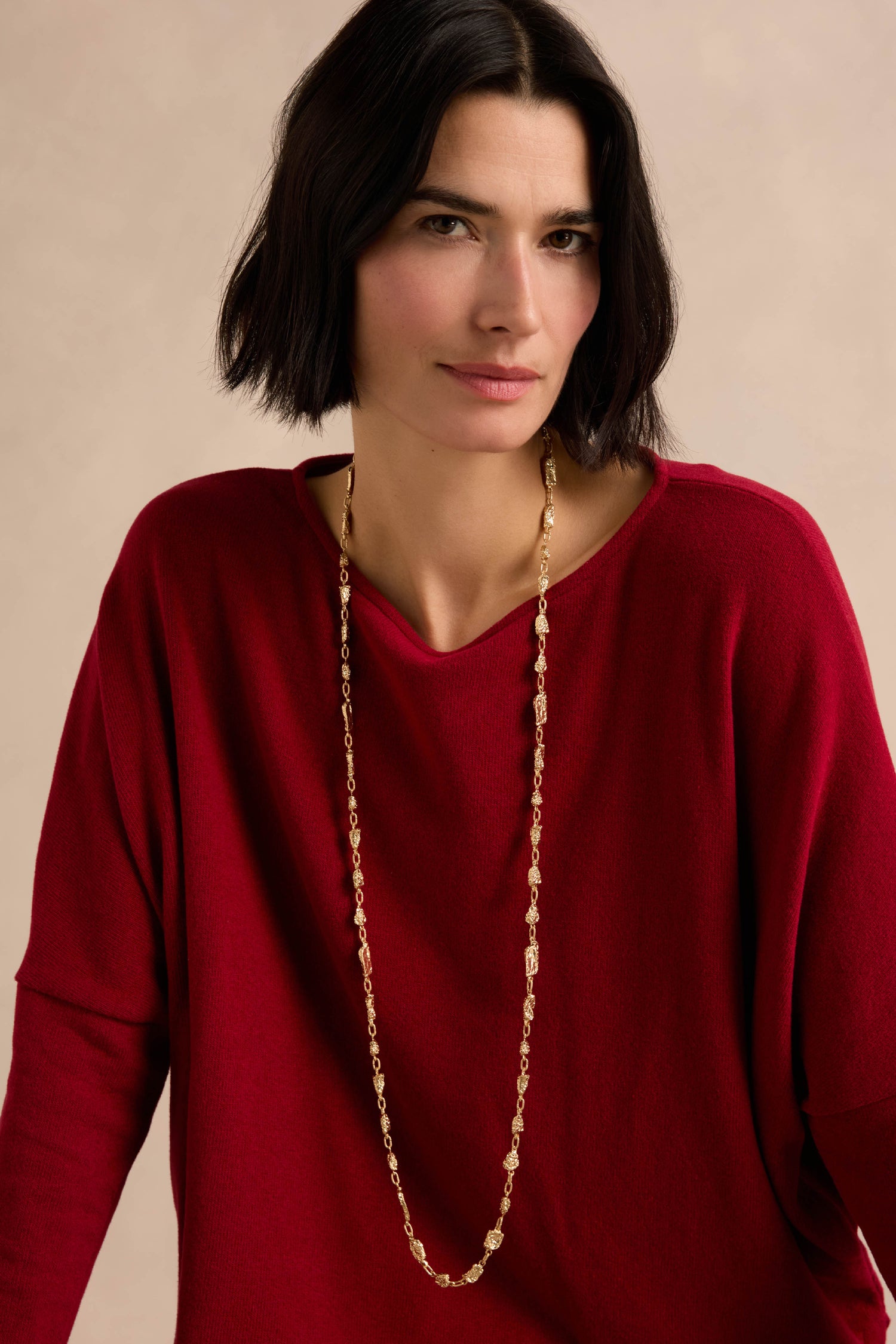 A woman with short black hair is wearing the Nugget Chain Necklace and a loose red top, standing against a neutral, textured surface.