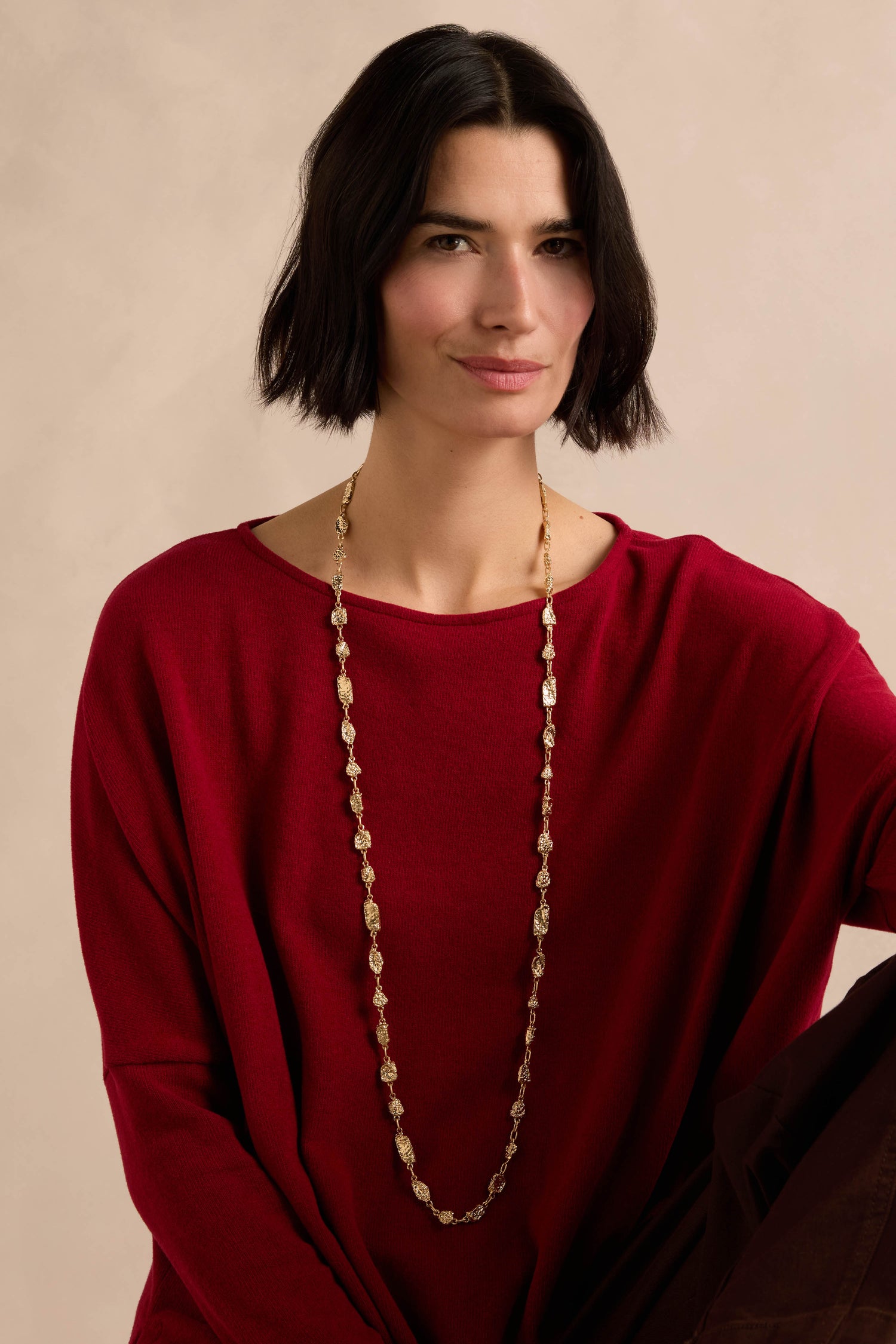 A woman with short dark hair sits against a textured surface, wearing a red top and the Nugget Chain Necklace.