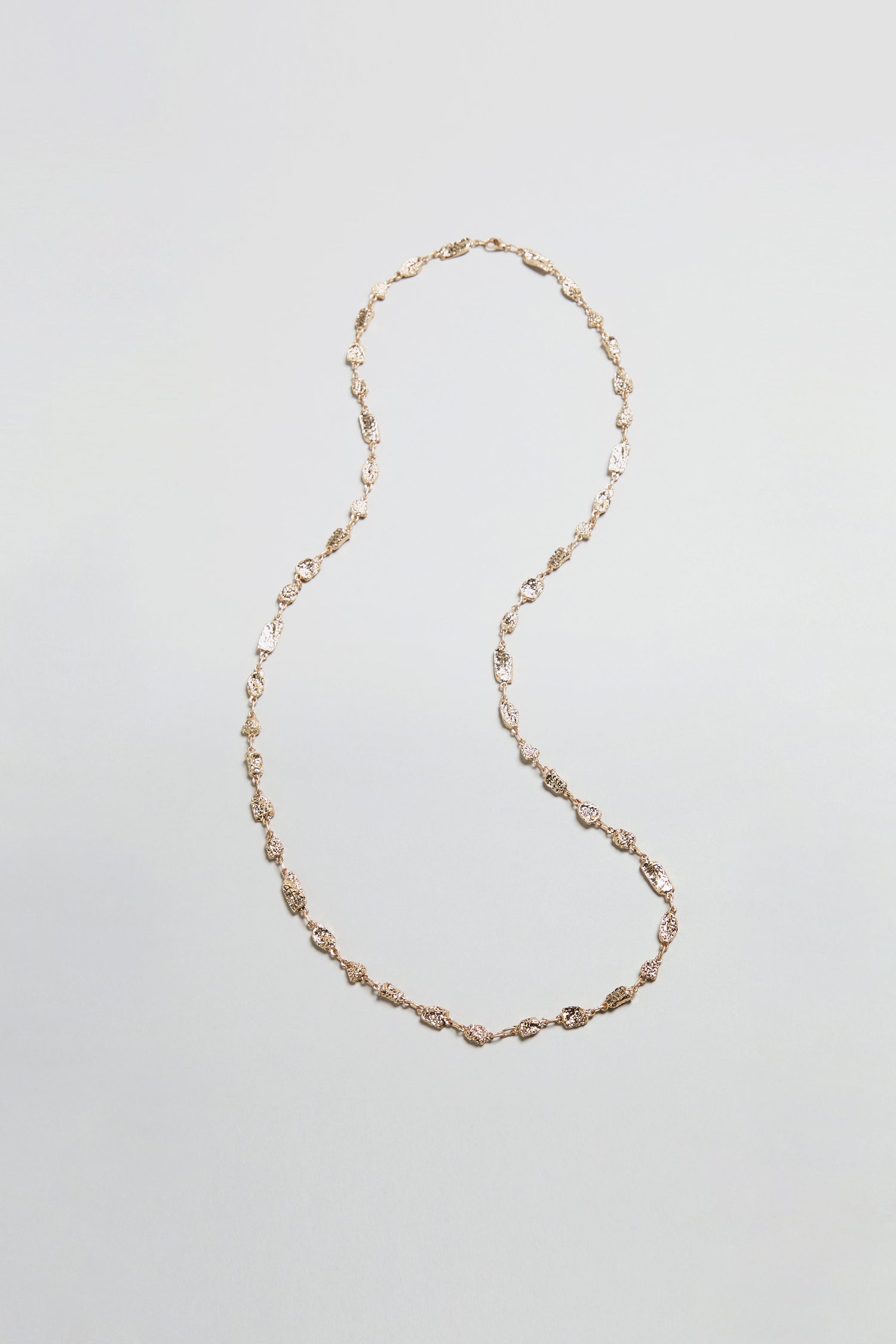 The Nugget Chain Necklace, featuring twisted links and a textured gold surface, is arranged in an S-shape on a plain, light gray background.