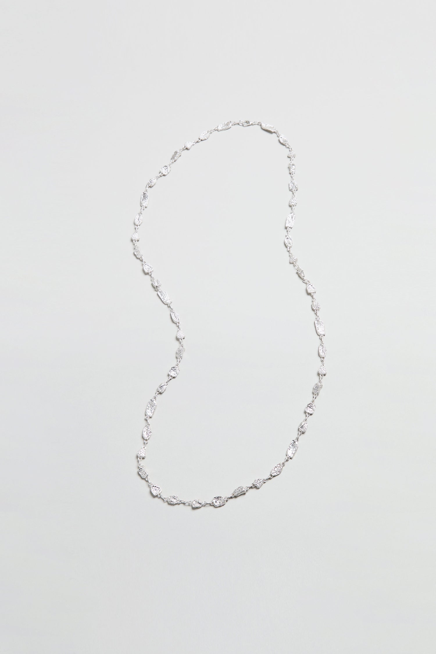 The Nugget Chain Necklace, featuring small, twisted silver links, is elegantly arranged in an S-shape on a plain, textured surface.