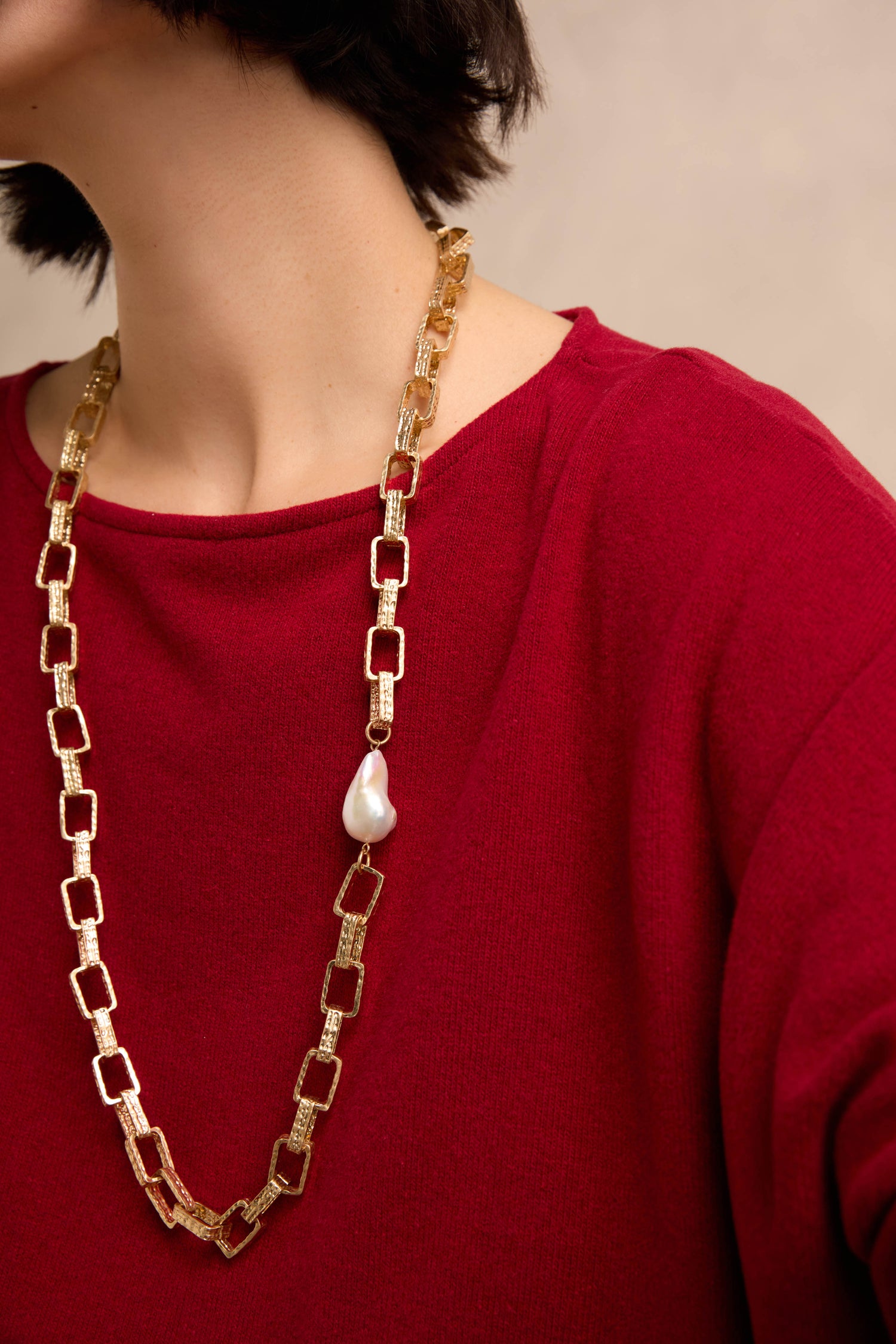 A person wearing the Gold Baroque Pearl Chain Necklace—a single large baroque pearl on a gold chain—paired with a red textured top; only the lower part of their face, neck, and upper torso are visible.