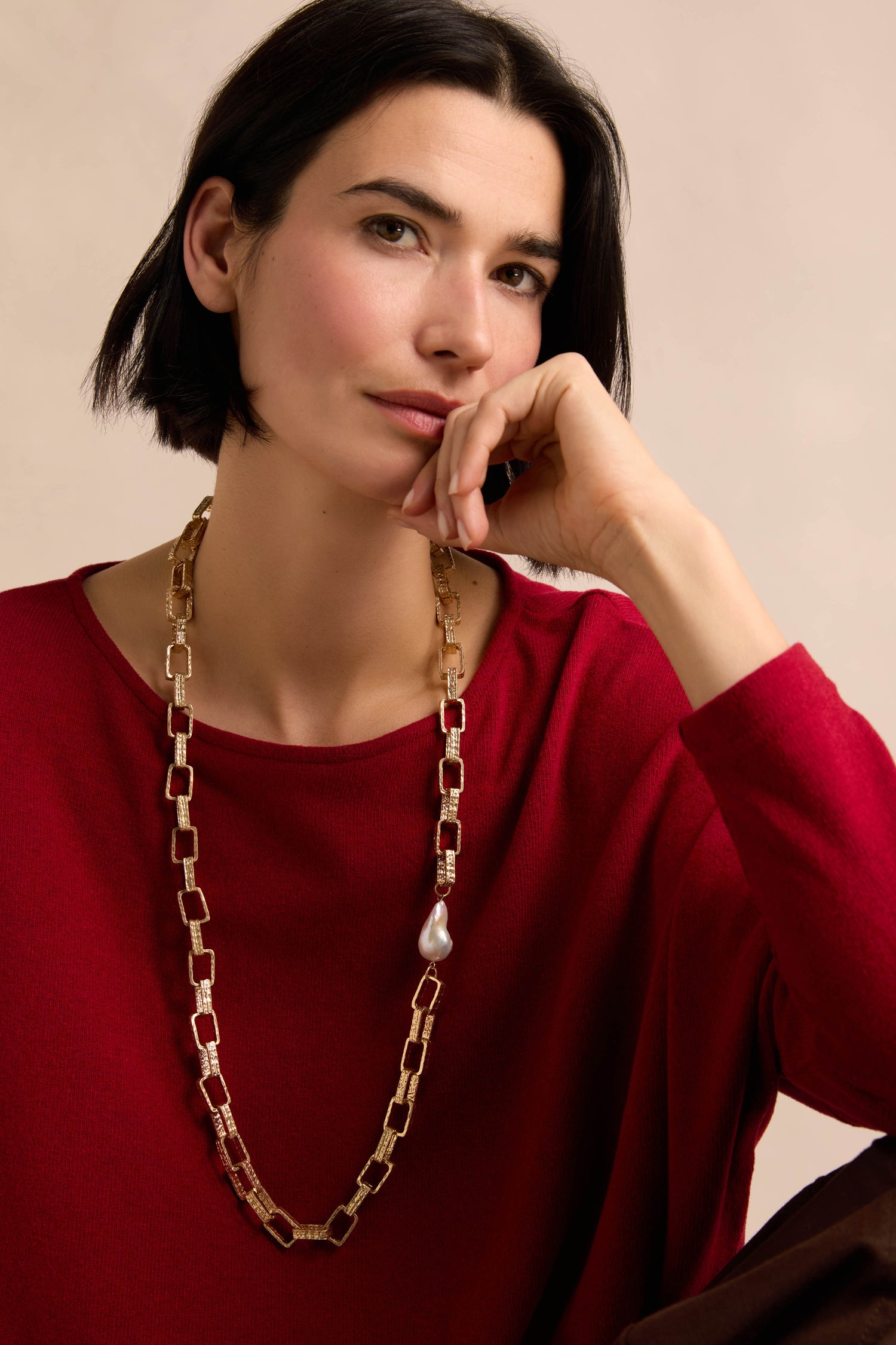 A woman with short dark hair, dressed in a red top, wears the Gold Baroque Pearl Chain Necklace—featuring a single pearl—as she rests her chin on her hand against a neutral background.