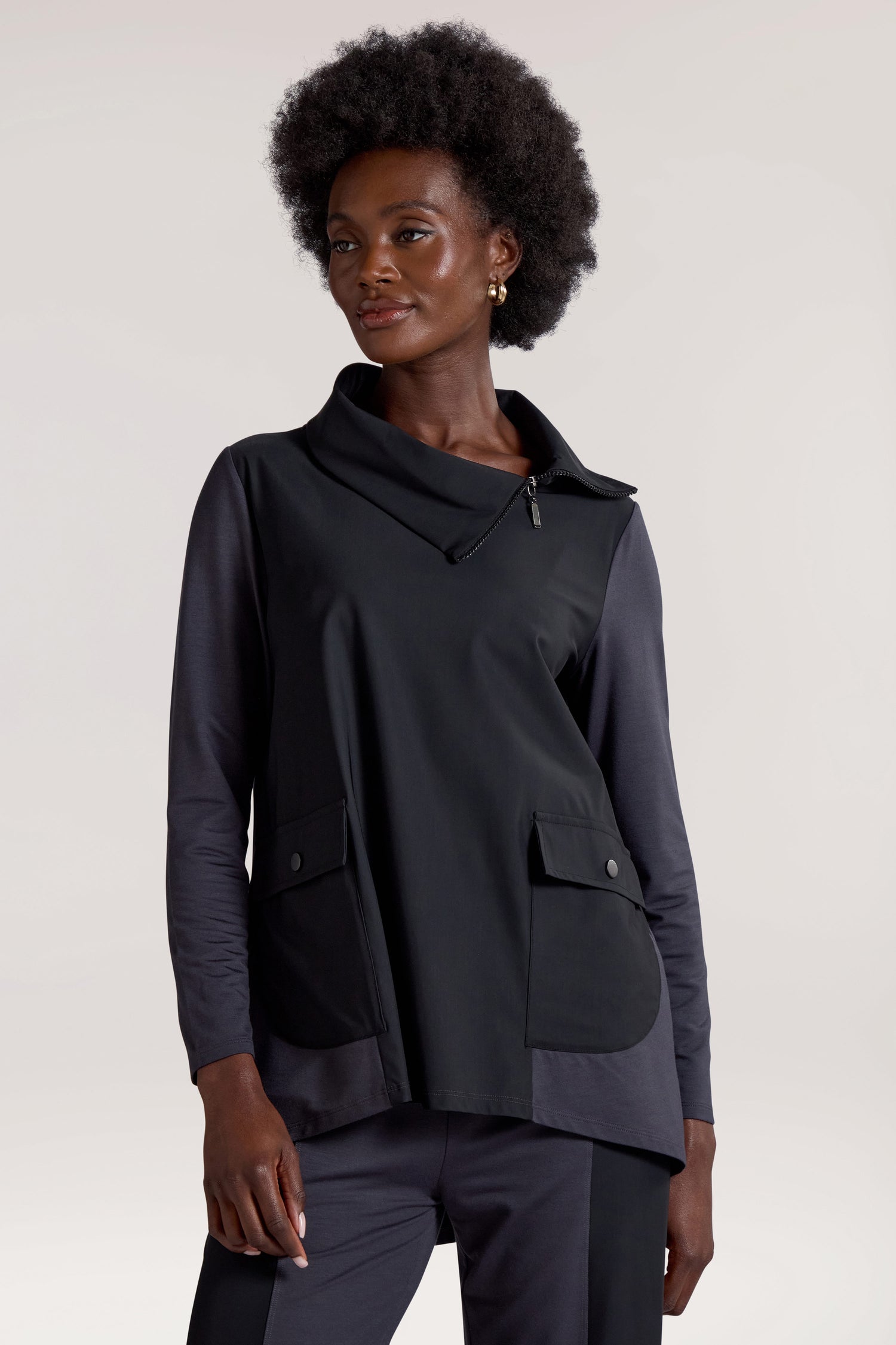 A person with an afro hairstyle stands confidently, sporting a Zip Collar Pocket Top in black, featuring a stylish zip collar and shawl detail. The top boasts two front pockets for convenience. The backdrop is a simple light color, keeping the focus on their chic ensemble.