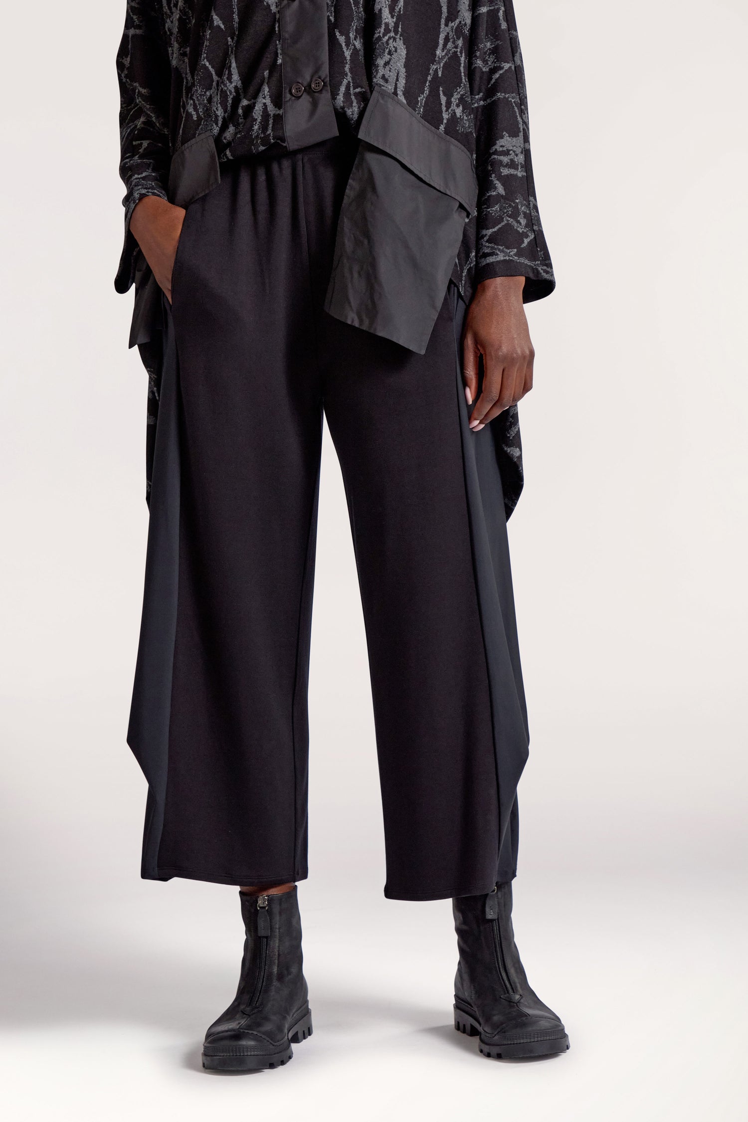 A person wearing Side Panel Pleat Hem Trousers with an elasticated waistband and wide legs, a black patterned top with an asymmetric hem, and black boots stands with hands in pockets. Only the torso and legs are visible.