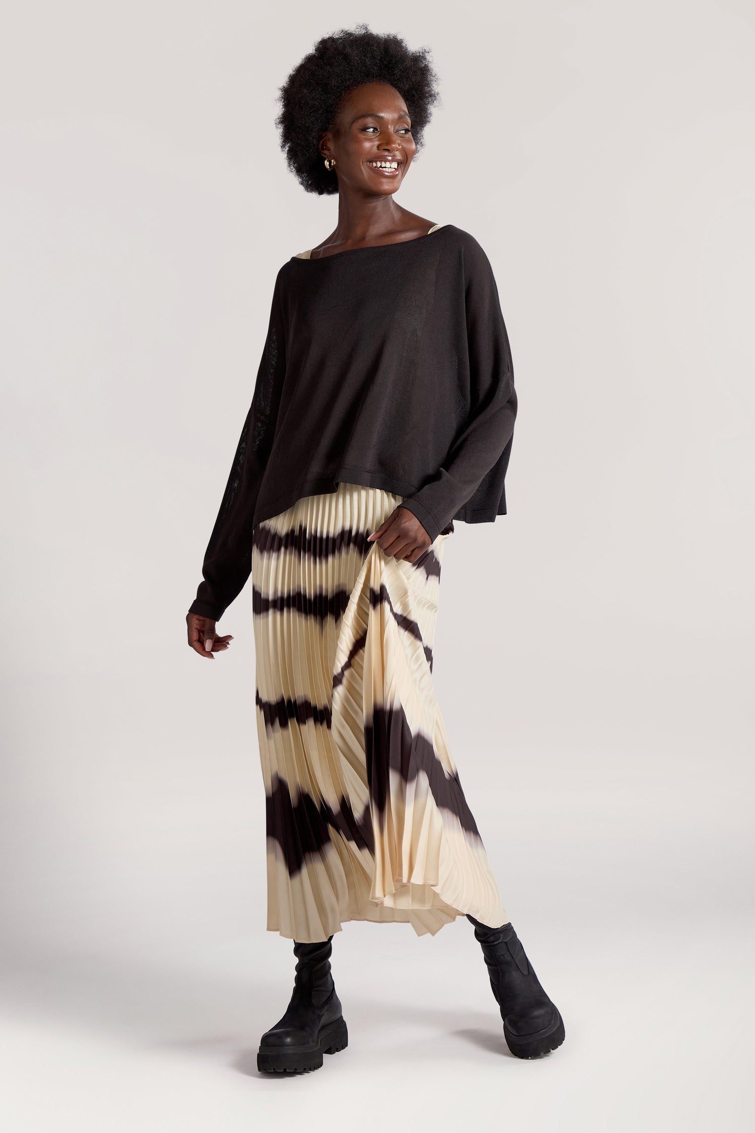 A person is standing and smiling, wearing the Tie Dye Pleat Dress with Jersey Top featuring black and beige pleats and accessorized with black boots.