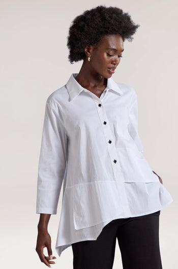 A person wearing the Panel Hem Shirt in white, featuring a wide collar and an asymmetric design that adds a unique touch, paired with black pants. They have short curly hair and are looking down with one hand in their pocket.