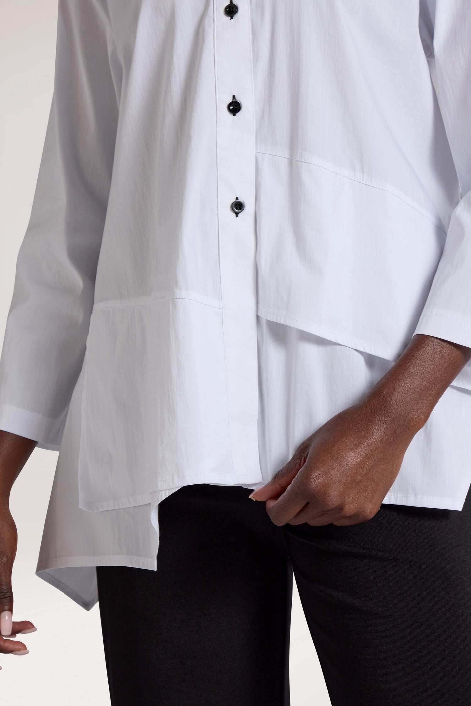 A person wearing a white Panel Hem Shirt — distinguished by its asymmetric design, black buttons, and three-quarter length sleeves — paired with black pants. The modern shirt adds a contemporary flair to the outfit.