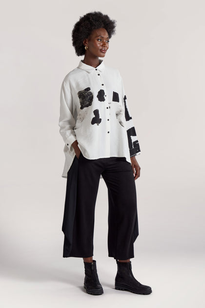 A woman stands posing, wearing the Viscose Print Easy Shirt with abstract black patterns, black wide-leg pants, and black ankle boots. She has an afro hairstyle and is looking off-camera to the side.