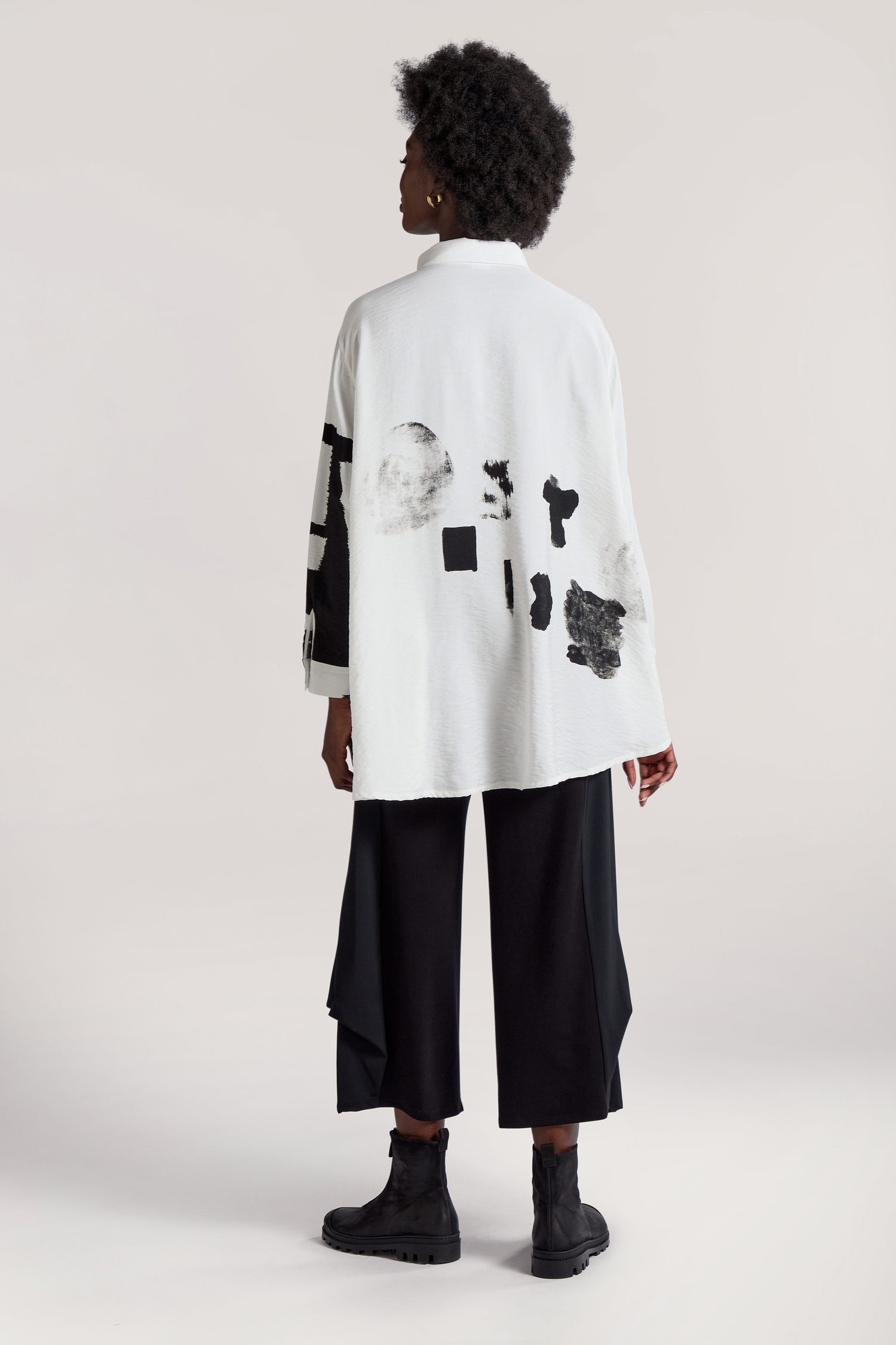 A person with an afro hairstyle is wearing the Viscose Print Easy Shirt, featuring white fabric adorned with black abstract designs, paired with black pants. Viewed from behind, this ensemble exudes a vibrant and modern visual in contemporary design.