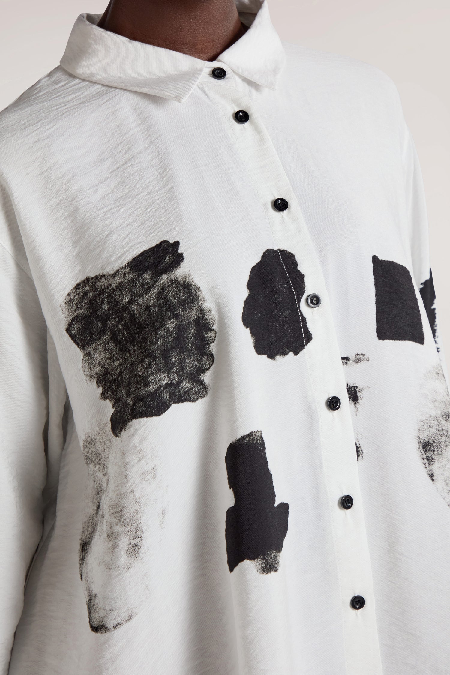 Close-up of a person wearing the Viscose Print Easy Shirt, a white button-up shirt adorned with a black, abstract paint pattern. This contemporary design features a collar and black buttons down the front.