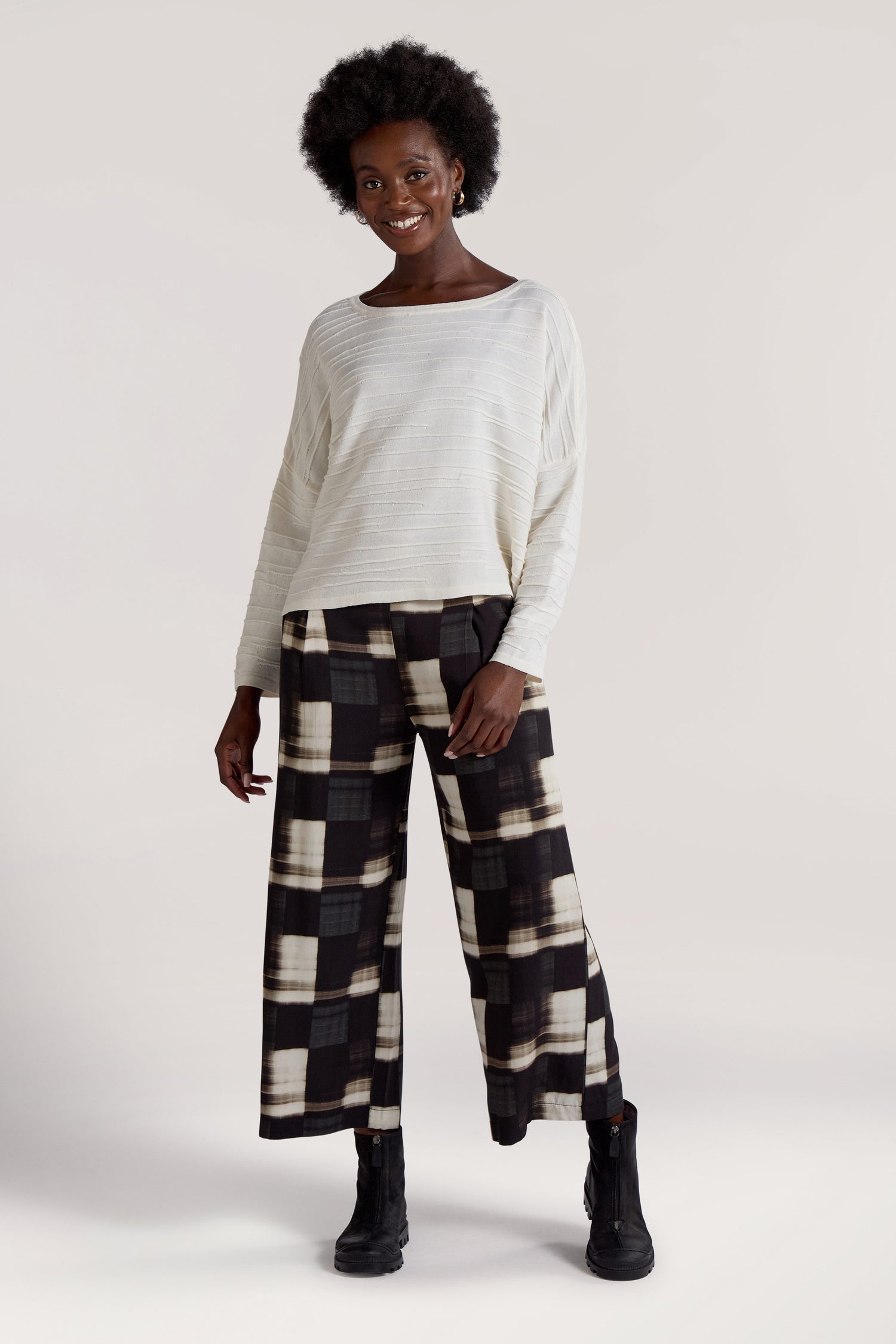 A woman with an afro hairstyle stands against a plain background, dressed in a long-sleeve white top paired with Blurred Check Trousers, featuring an elasticated waistband. Her look is completed with black lace-up boots. She is smiling.