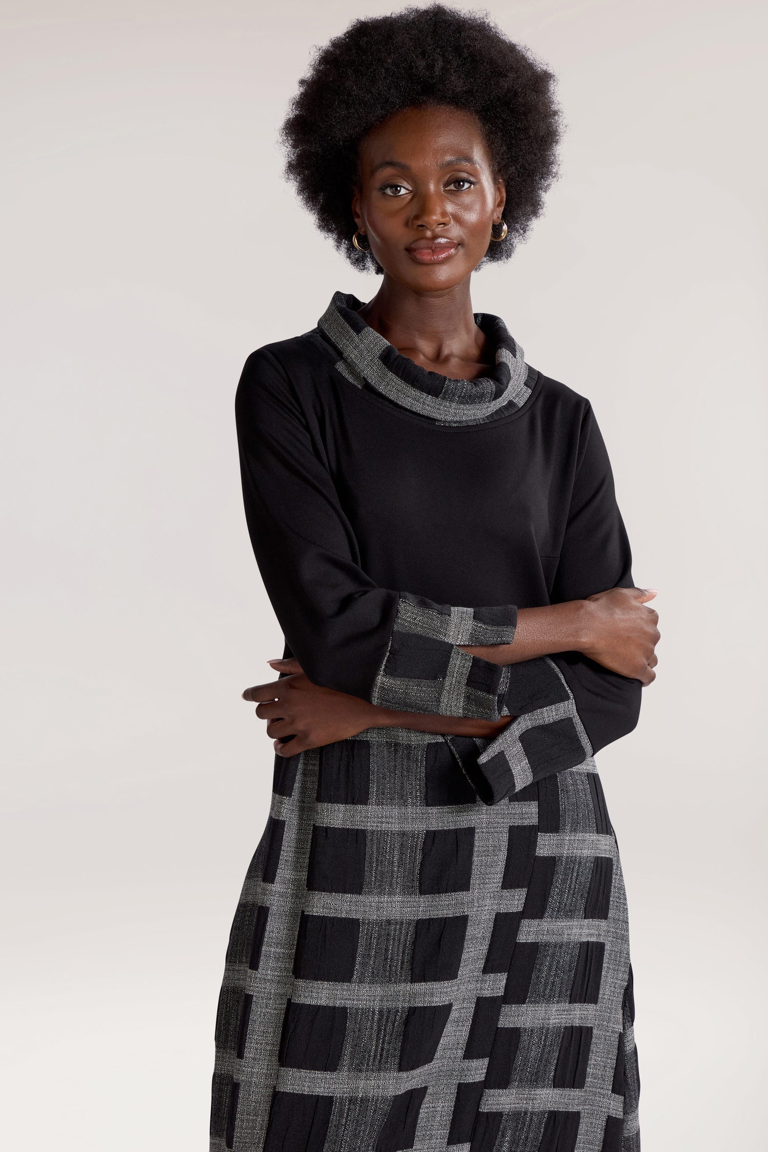 A person with an afro hairstyle wearing a Checked Cowl Neck Dress featuring a black check print and grey checkered patterns on the lower part and sleeves stands with arms crossed against a neutral background.