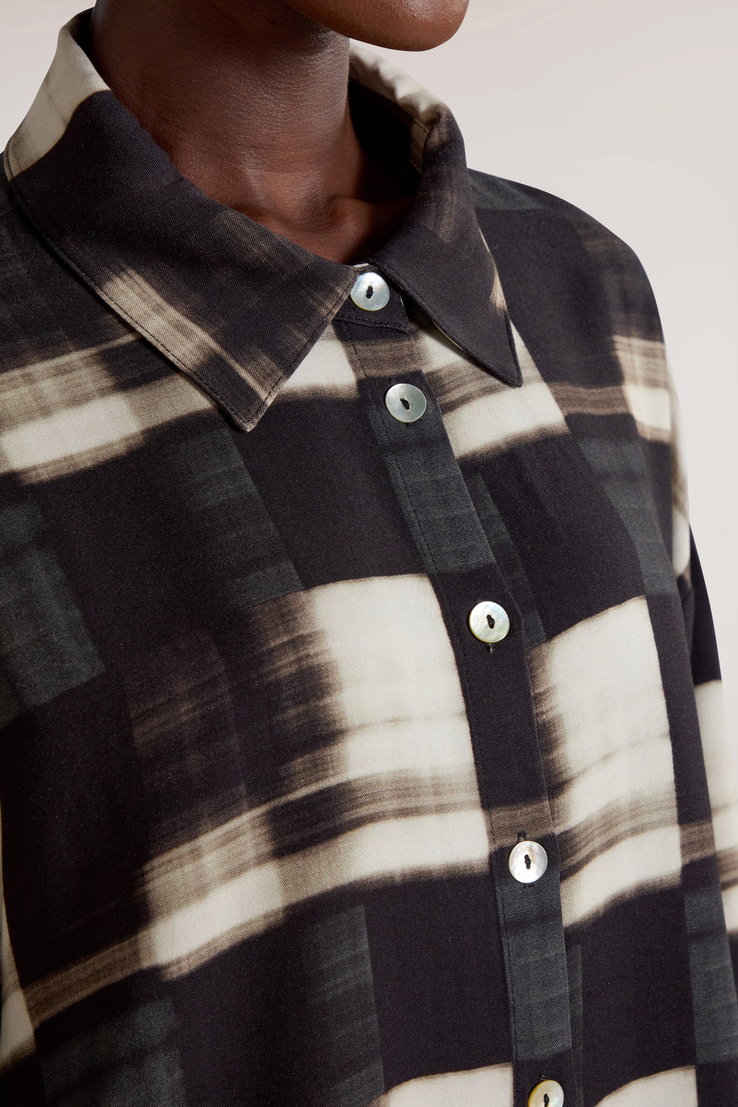 Close-up of a person wearing the Blurred Check Boxy Shirt, which features a black and white plaid pattern with large buttons, highlighting its minimalist design and contemporary flair. Only the neck and upper torso are visible.