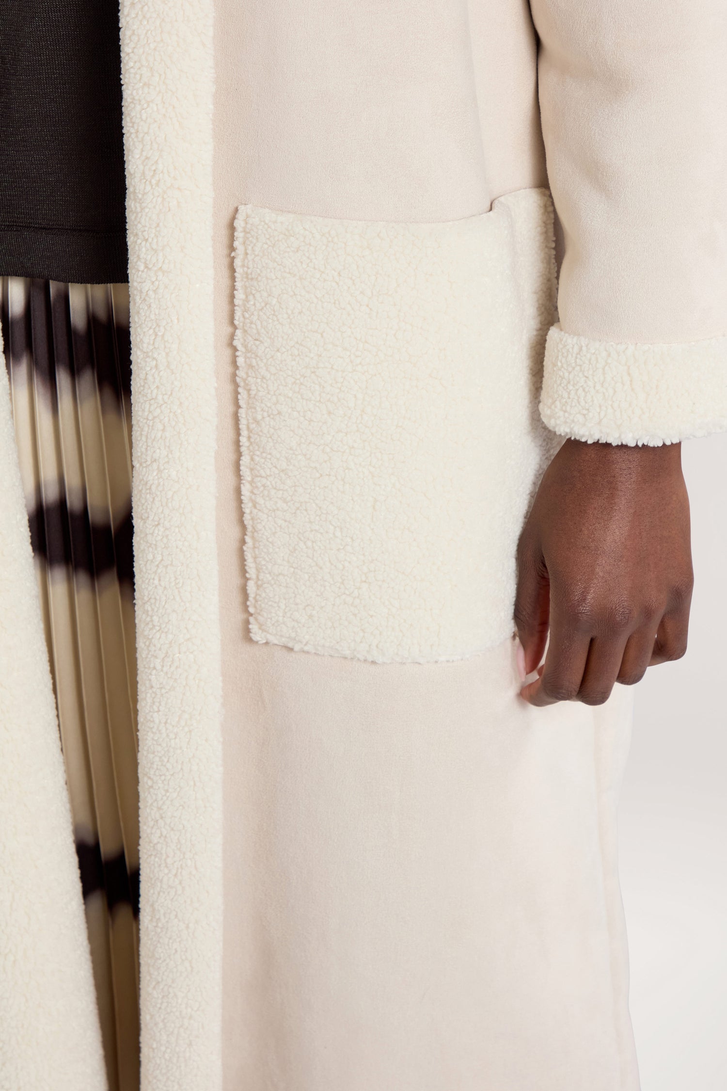 Close-up of a person wearing a cream-colored Faux Sheepskin Coat featuring a large, textured front pocket. The person's hand rests by their side. The coat is paired with a black and white patterned skirt, creating a modern minimalist look.