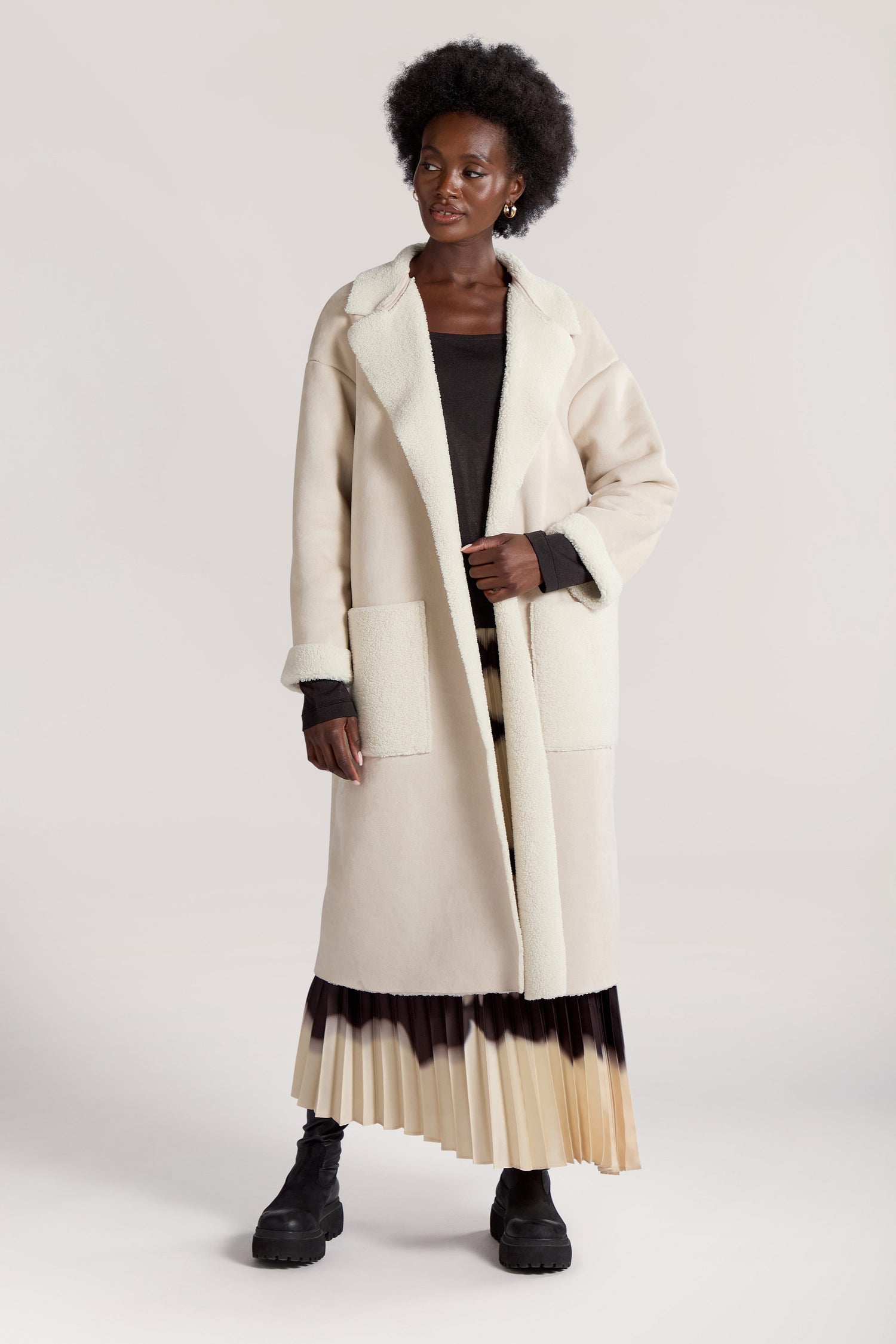 A person is standing against a neutral background, wearing a long cream-colored Faux Sheepskin Coat with two front pockets, a black top, a pleated skirt with a gradient pattern, and black boots.