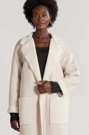 A person dressed in a long-line faux sheepskin coat, named the Faux Sheepskin Coat, stands against a plain background. The light-colored coat complements their black top and features large front pockets and collars, exuding a modern minimalist vibe.