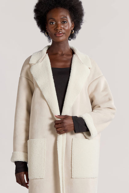 A person dressed in a long-line faux sheepskin coat, named the Faux Sheepskin Coat, stands against a plain background. The light-colored coat complements their black top and features large front pockets and collars, exuding a modern minimalist vibe.