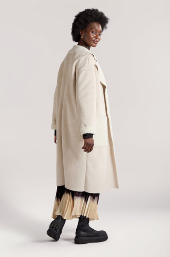 A woman is wearing a long-line length Faux Sheepskin Coat over a pleated skirt and black boots, standing and smiling against a plain background, capturing the essence of modern minimalist style.