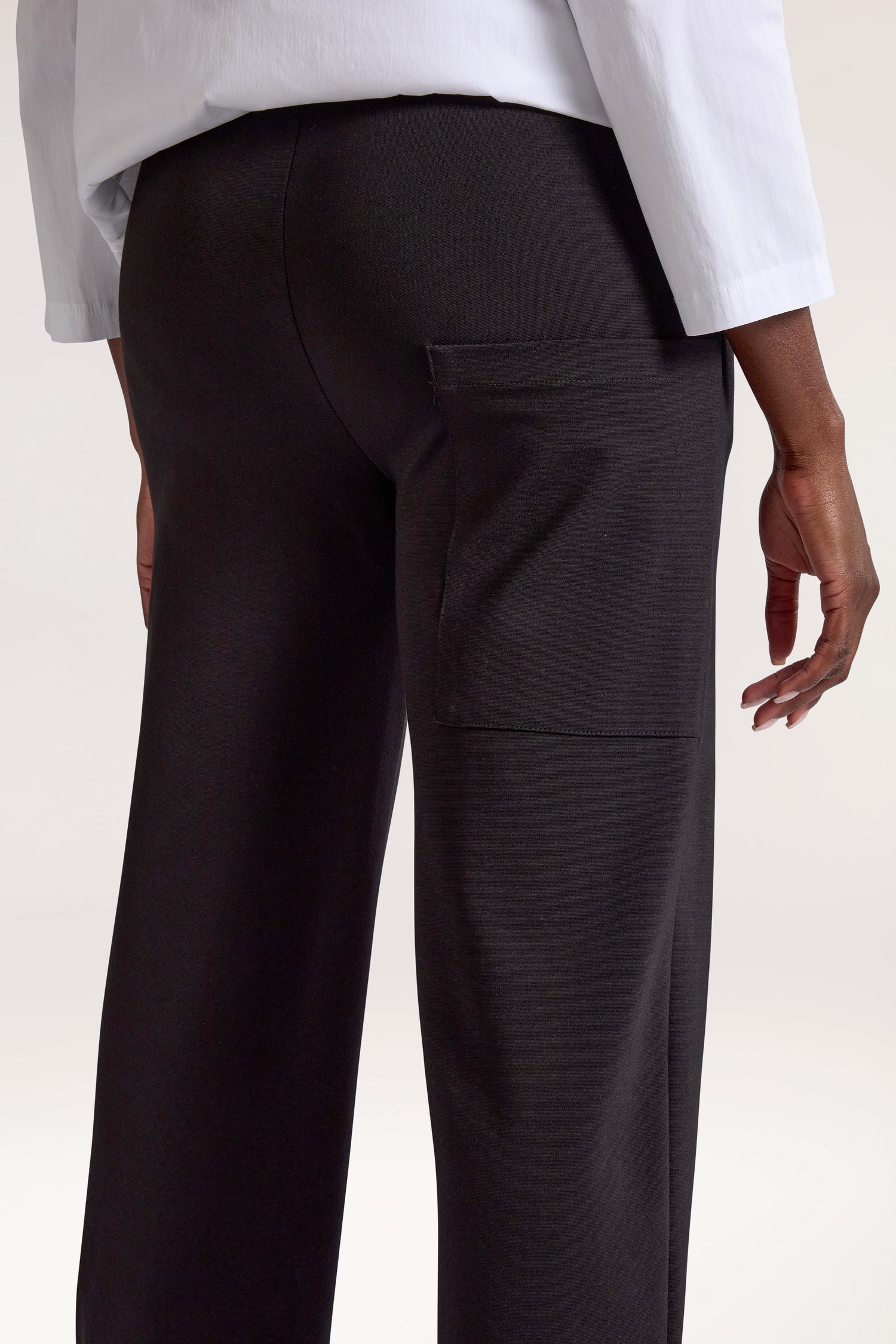 A person wearing the black Ponte Cuff Hem Trouser, featuring an elasticated waistband and a side pocket, seen from the back, paired with a crisp white shirt.