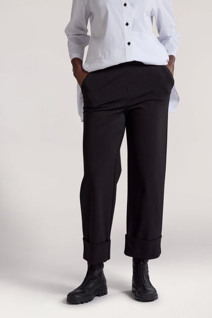 Person wearing a white button-up shirt and black Ponte Cuff Hem Trouser with hands in pockets, paired with black ankle boots. The minimalist approach of the outfit highlights its sleek design and the comfort of the elasticated waistband. The image shows the outfit from neck to feet.