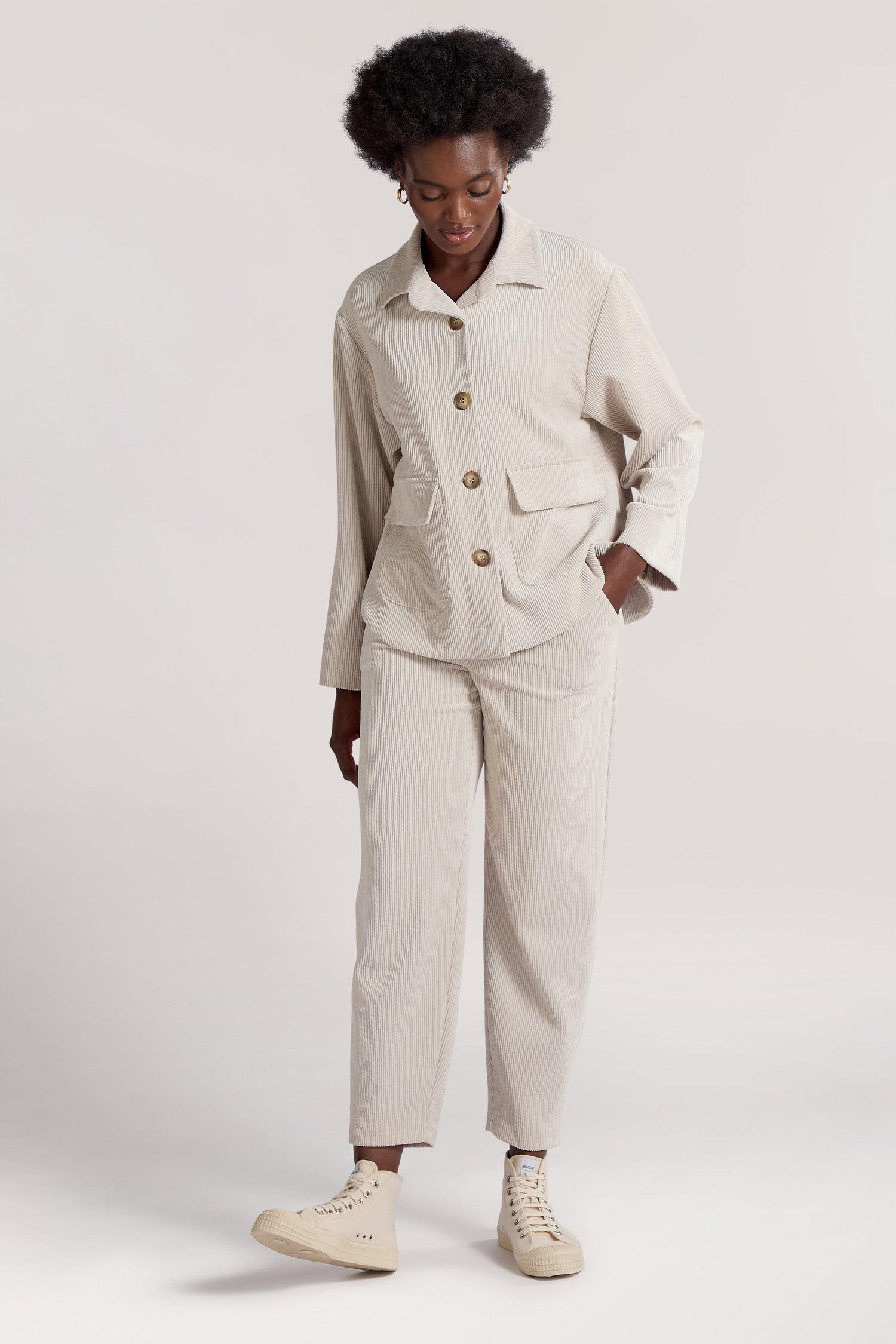 Stands against a plain background, a person wearing our light beige Cord Trouser and matching long-sleeve buttoned jacket with front pockets. They have one hand in their pocket and are sporting light-colored sneakers. The outfit is crafted from premium corduroy fabric.