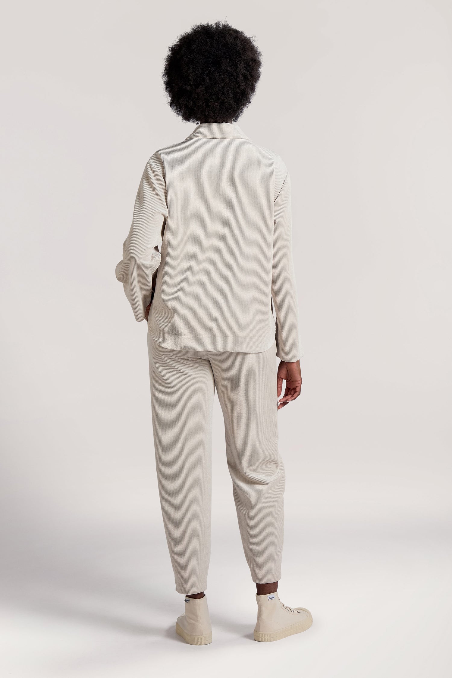 The person with short curly hair is standing and facing away, wearing the Cord Jacket in a light color along with matching pants, both showcasing a minimalist appeal.