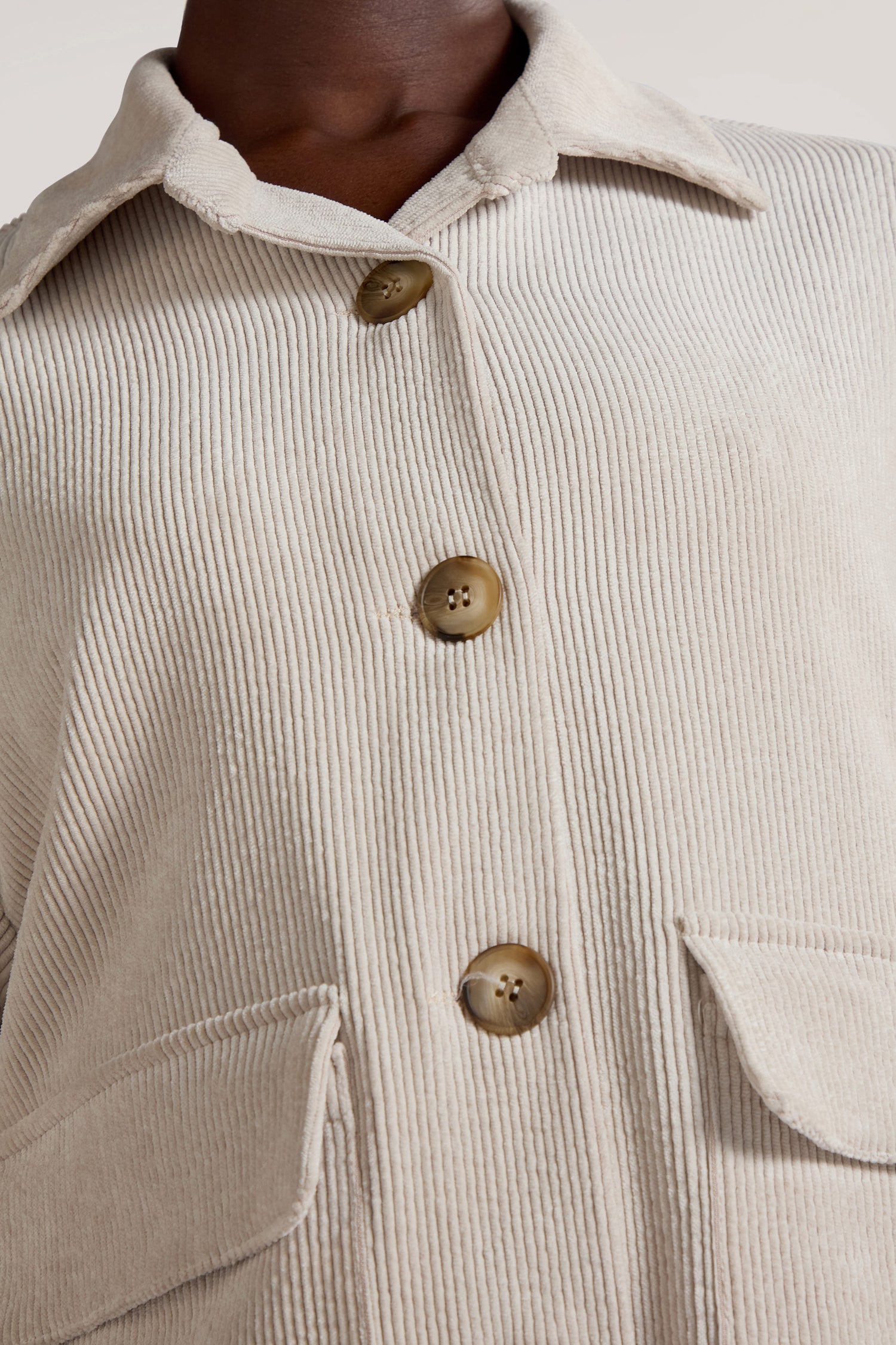 Close-up of a person wearing the Cord Jacket, a beige piece with large brown buttons and two front flap pockets, showcasing the minimalist appeal of its corduroy fabric.