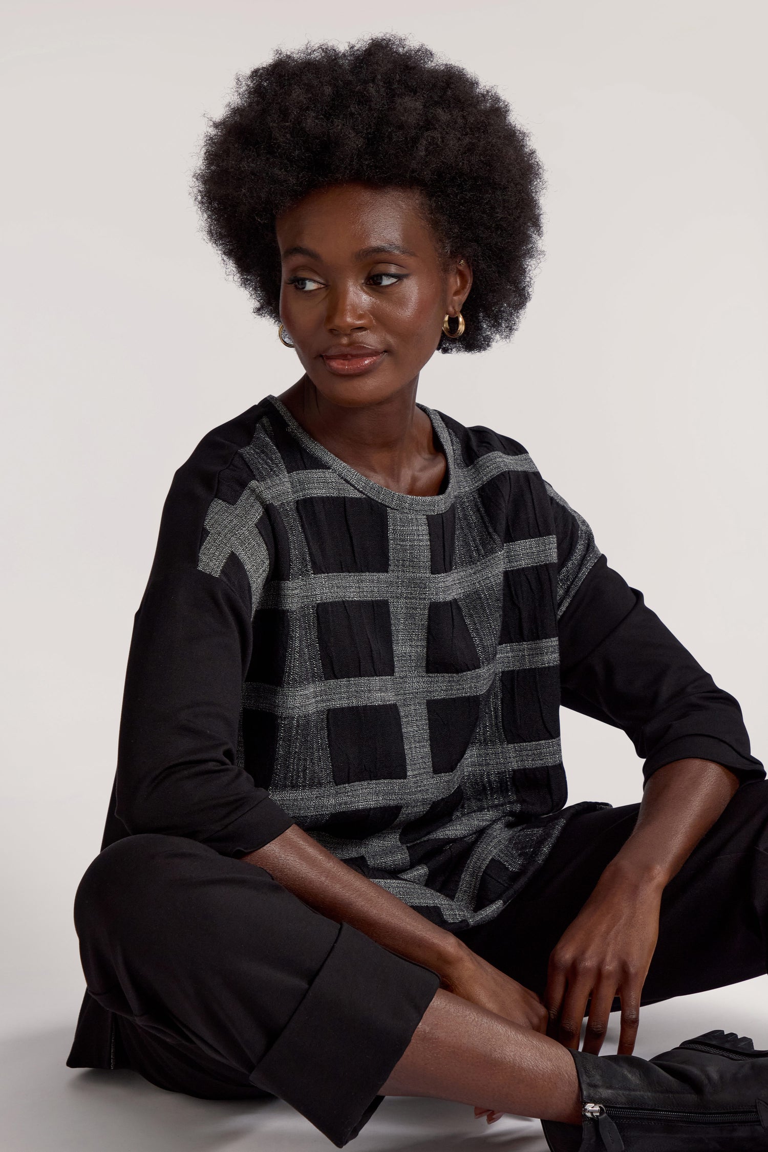 A person with natural hair, exuding relaxed elegance in a Checked Tunic in black and gray paired with black pants, is sitting against a plain background.