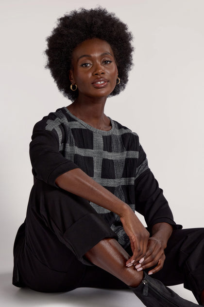 A woman with short curly hair sits on the floor, radiating relaxed elegance in her Checked Tunic and black pants. She gazes at the camera with a neutral expression, embodying contemporary design.